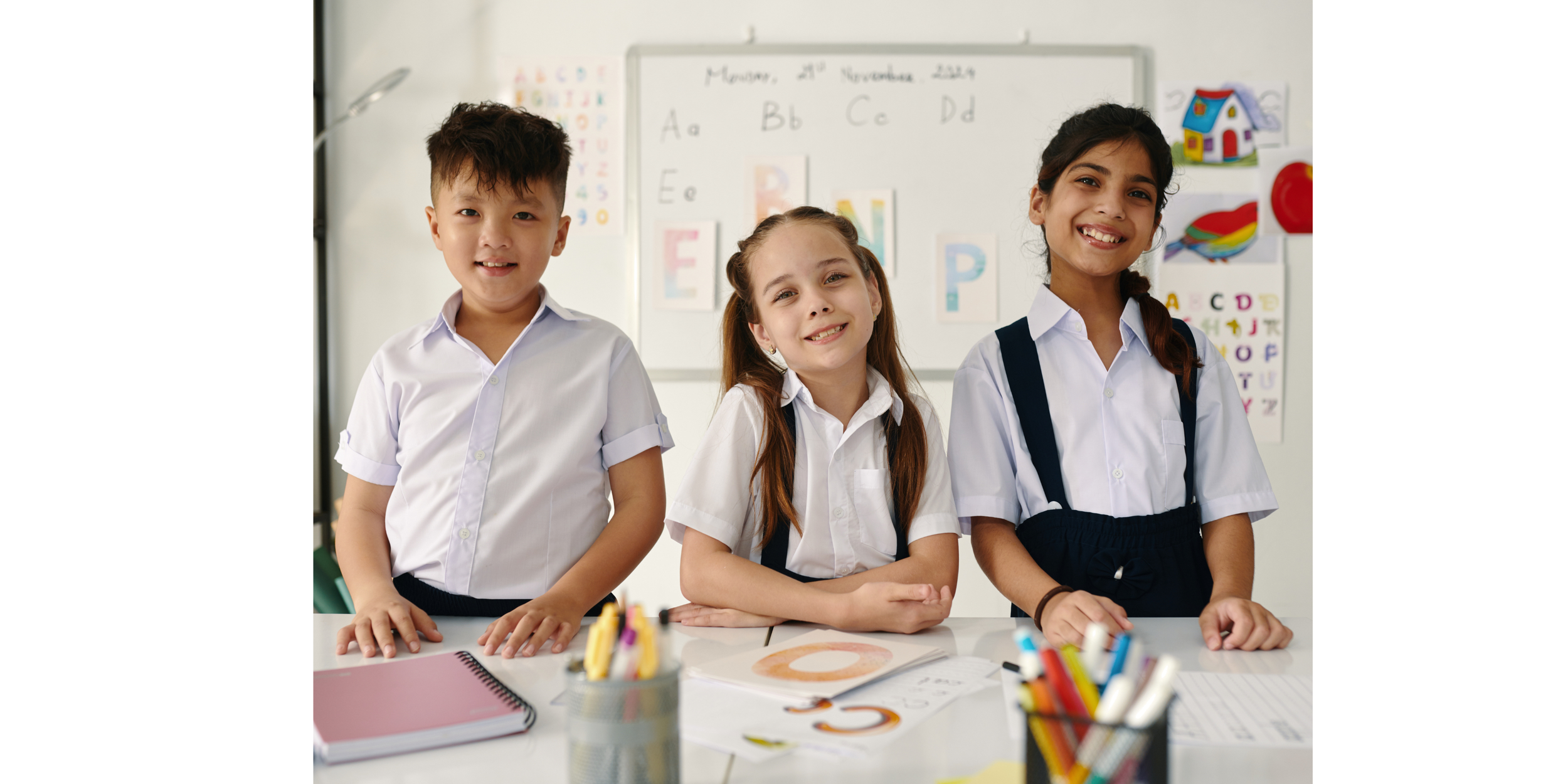 Three school children in white uniforms smiling at the camera in a classroom with educational posters and materials in the background.