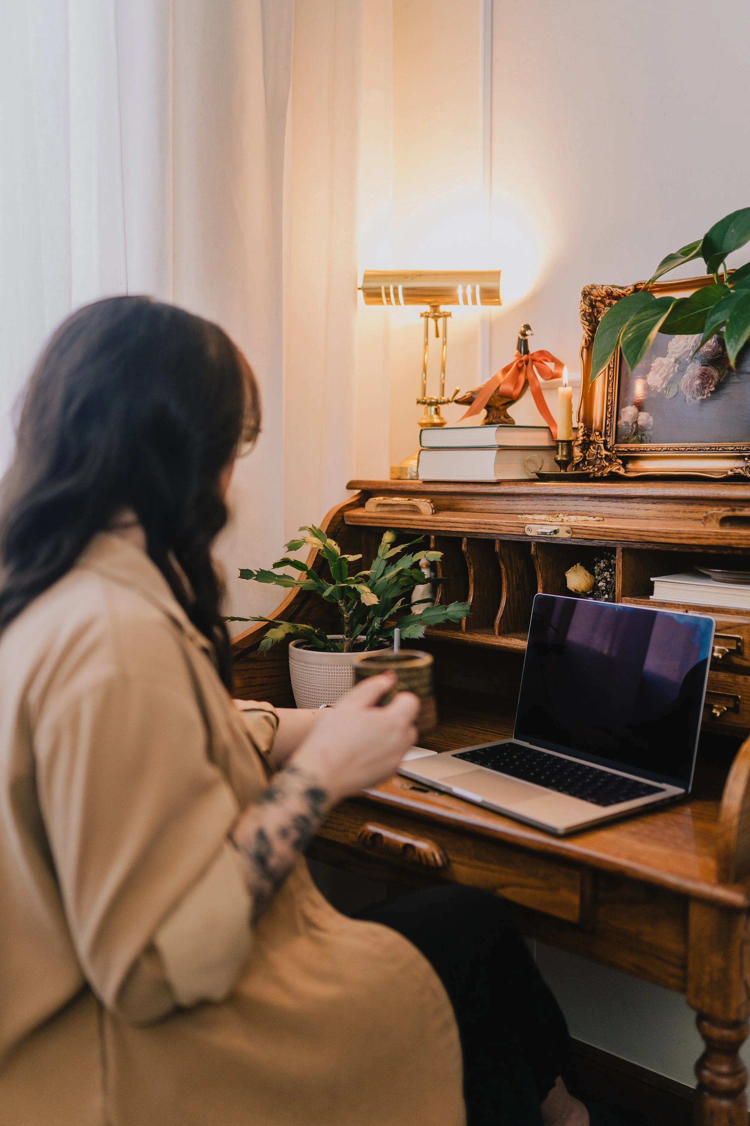 Joanna Jackson, LPC, sitting in front of a laptop at a roll top desk, holding a coffee mug. Her desk is decorated with plants, vintage items, and a goose statue.
