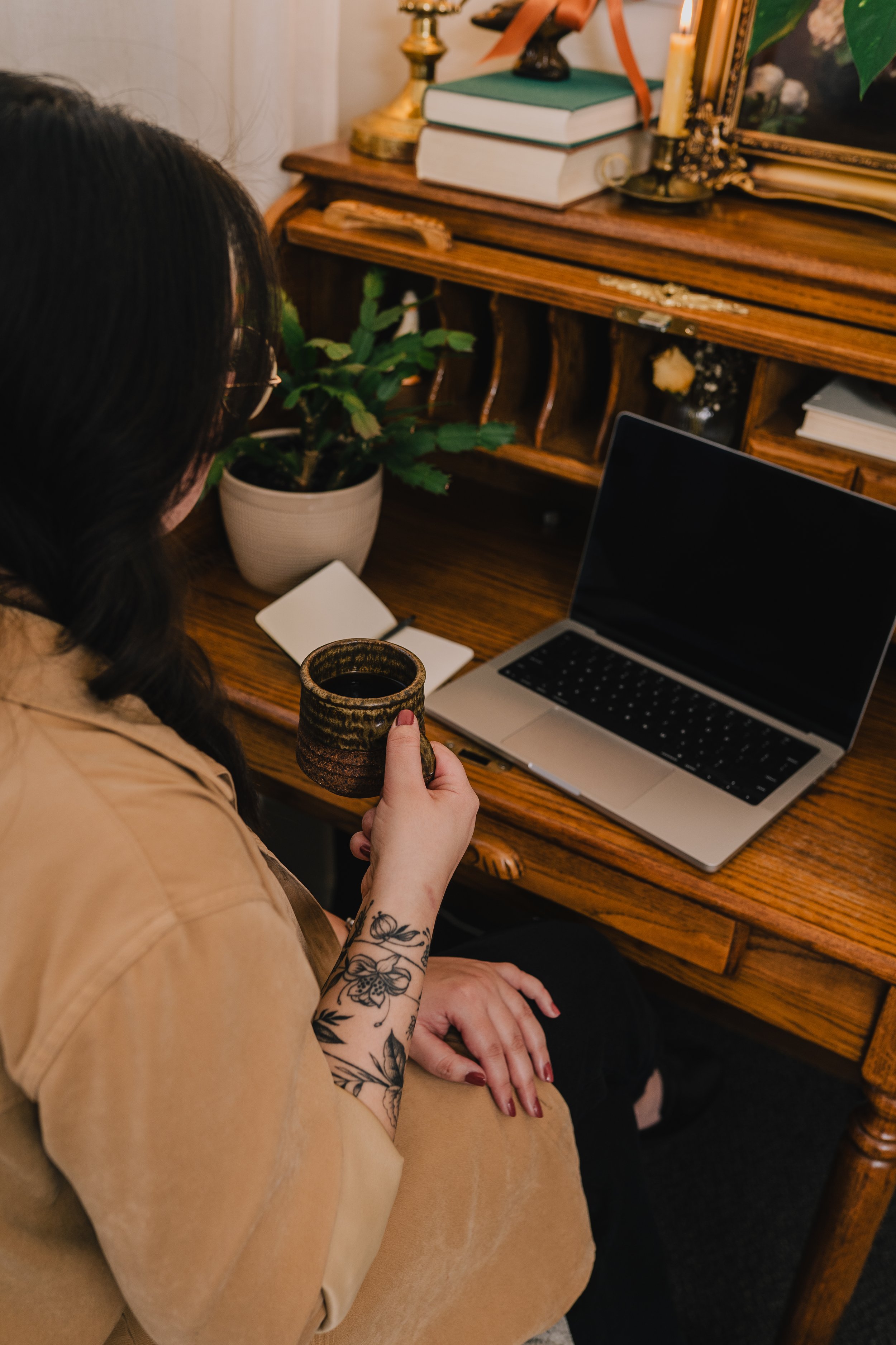 Joanna Jackson, LPC, sitting at her desk with a notebook and laptop in front of her. She's holding a coffee mug and her desk is decorated with books, plants and vintage items.