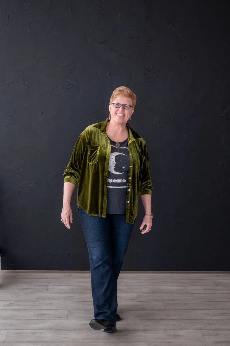 Full-length portrait of Susan Lynn smiling in a green velvet shirt and jeans against a dark studio background, representing grounded and approachable spiritual guidance.