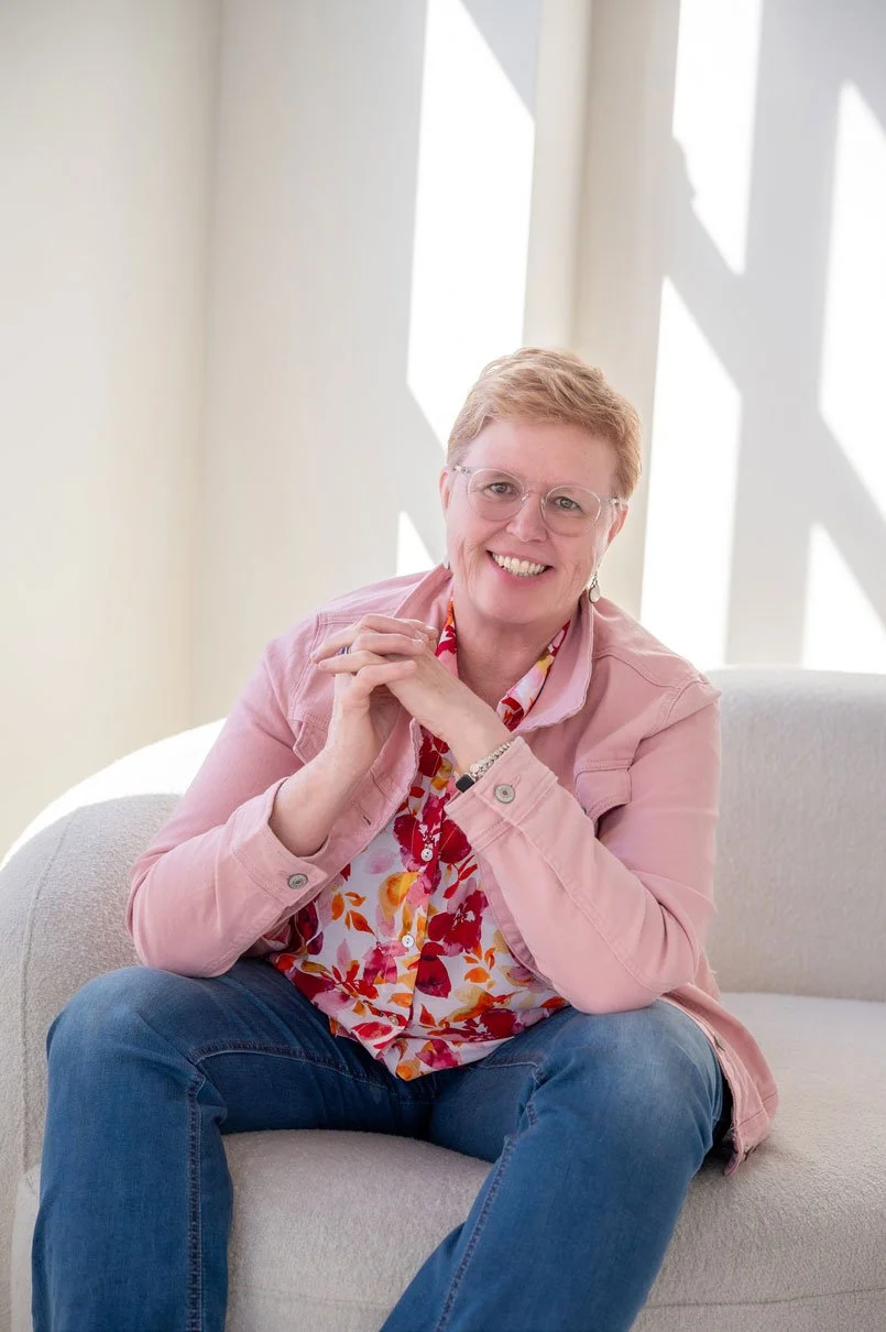 Susan Lynn smiling while seated in a chair, wearing a pink jacket and floral shirt, representing approachable and grounded spiritual guidance.