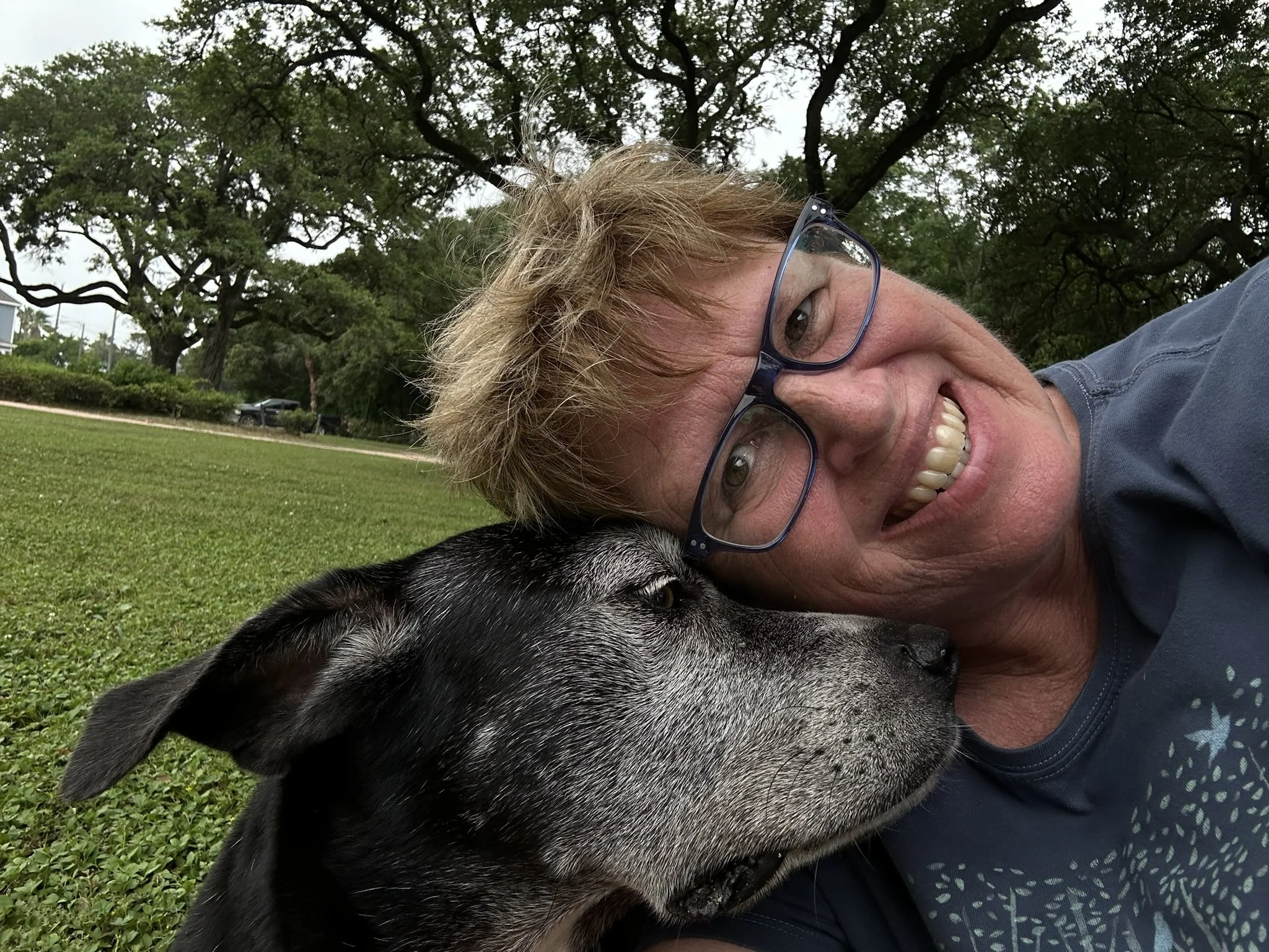 Susan Lynn smiling outdoors beside her black dog in a grassy park, showing her warm and approachable personality.