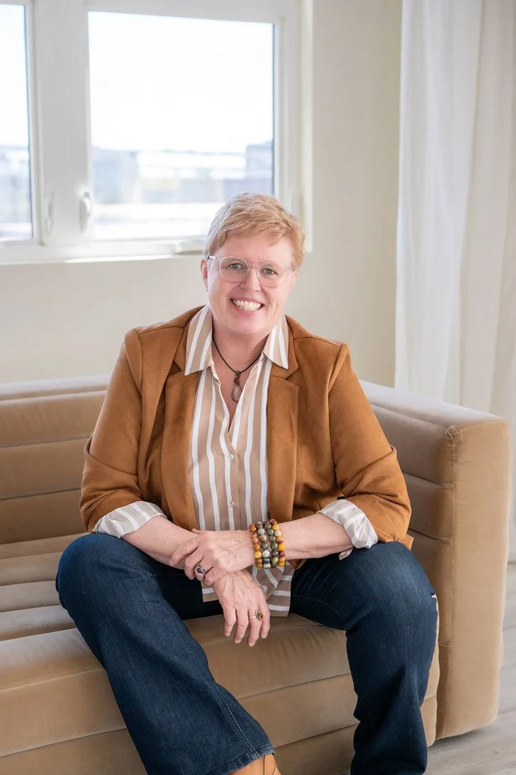Susan Lynn smiling while seated on a sofa, wearing a tan blazer and striped shirt, representing grounded spiritual guidance and intuitive teaching.