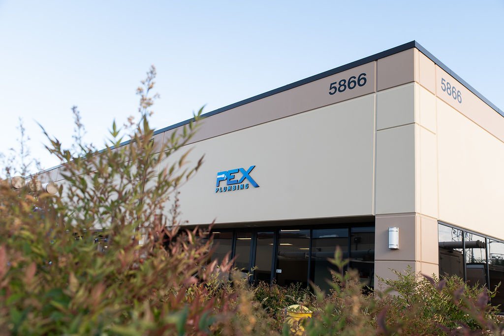 Exterior of a building with a sign that reads PEX Plumbing and the address 5866, with bushes in the foreground and a clear sky above.