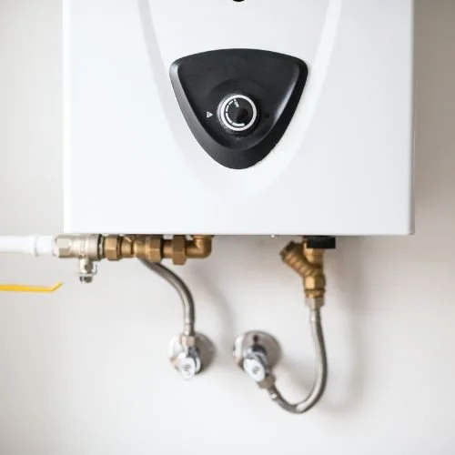 Close-up of a white tankless water heater with a black control dial on the front, connected to copper and metal pipes.