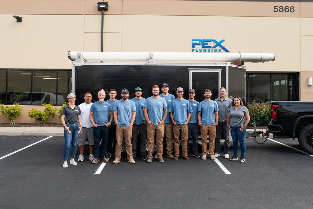 Group of 12 people standing in front of a black plumbing trailer with a large pipe on top, in a parking lot outside a building with a sign that reads 'PEX Plumbing'.