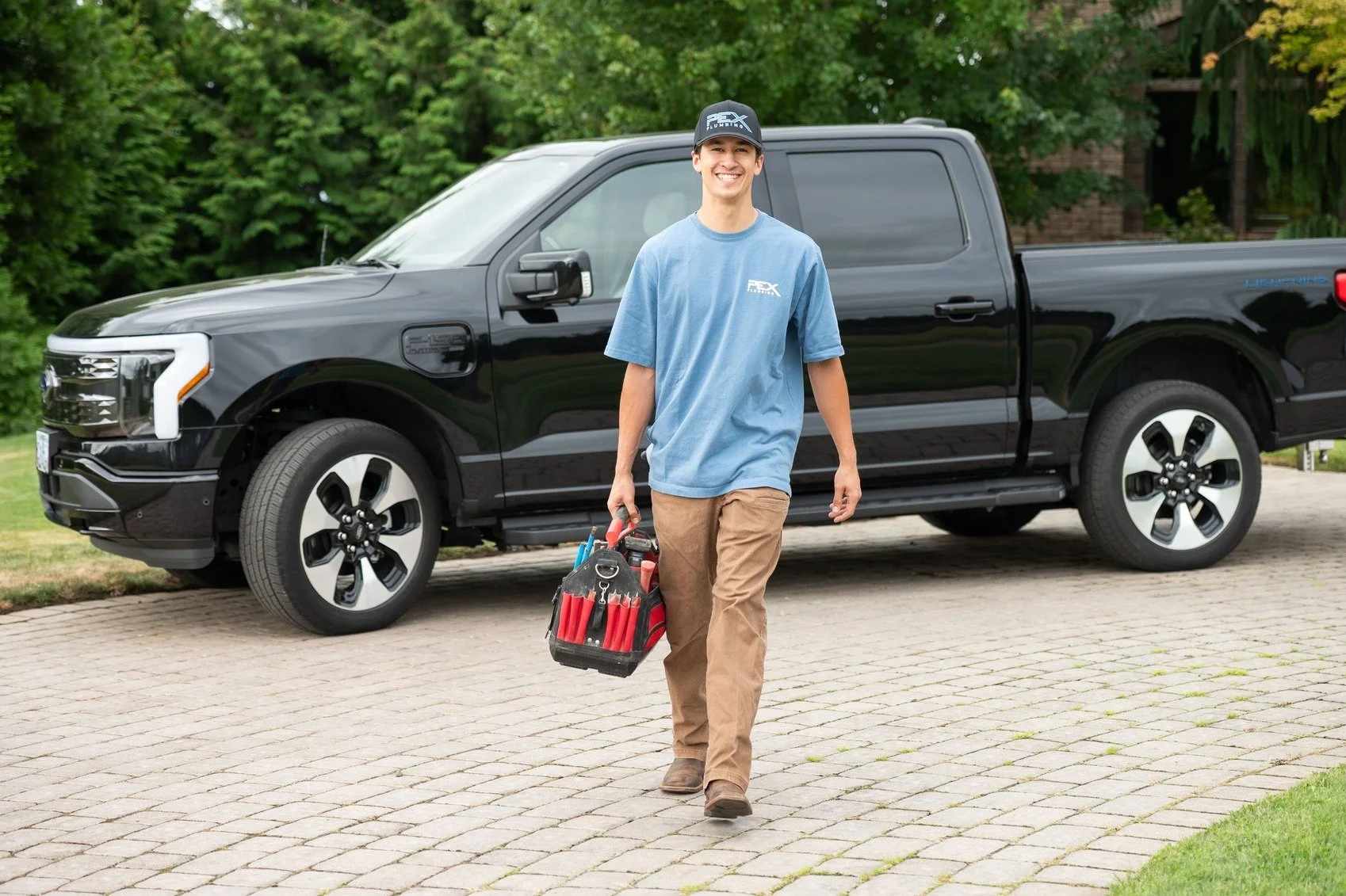 A young man in blue T-shirt and brown pants holding a black and red tool bag, walking on a brick driveway near a black pickup truck, with green trees in the background.