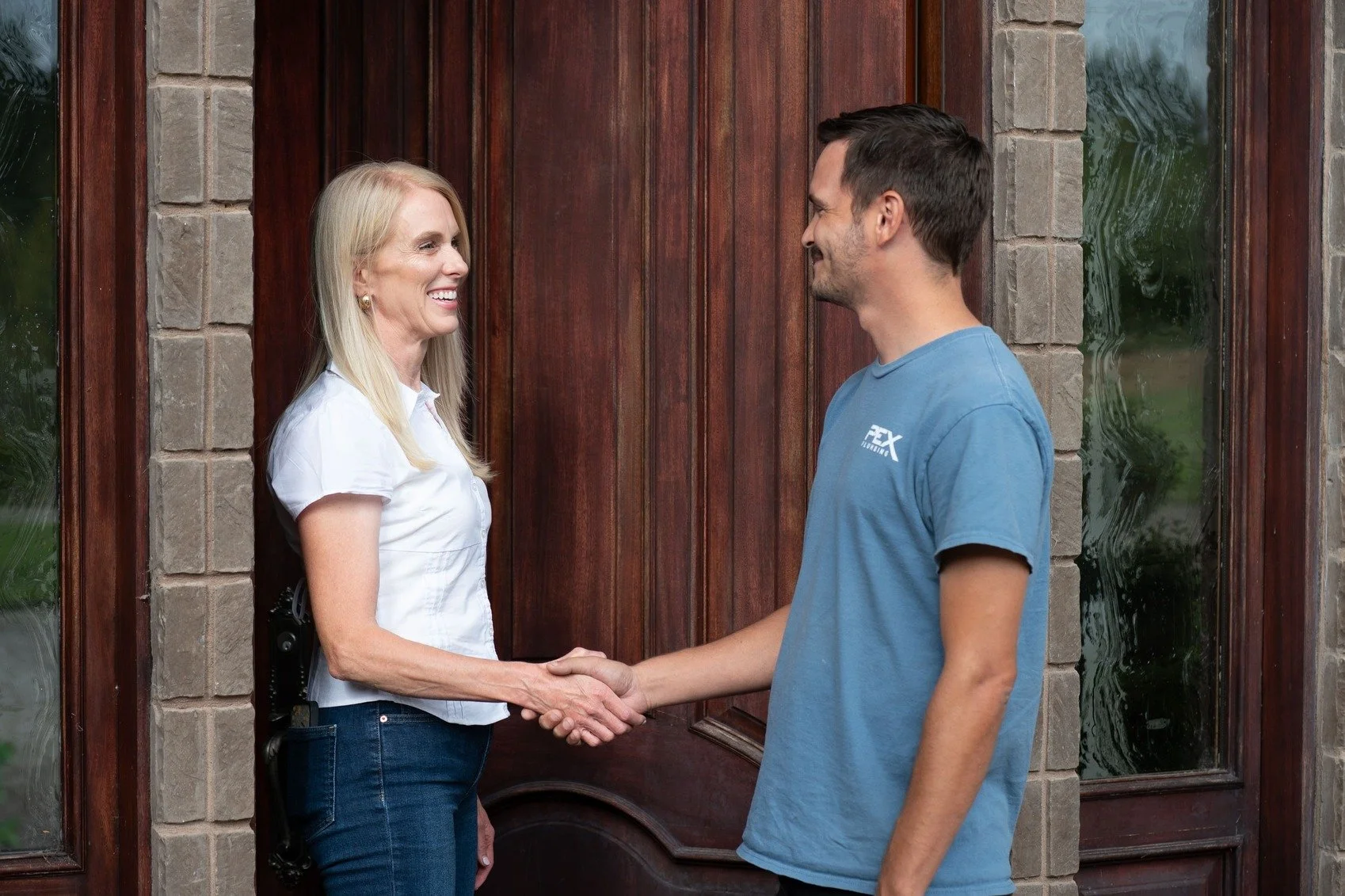 A woman and a man shake hands outside a house with a wooden door, smiling at each other.