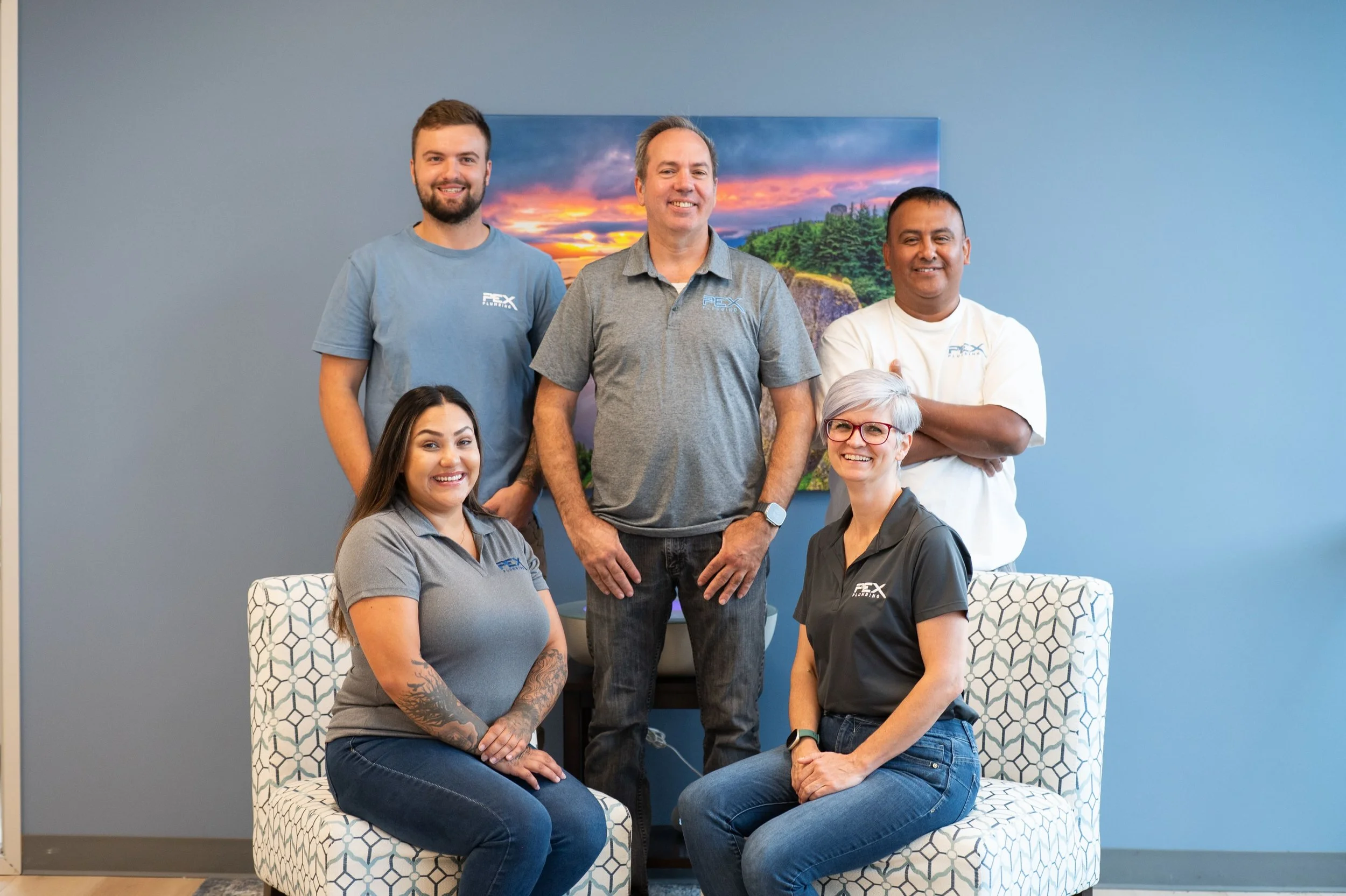 Group of six people smiling in an office setting, two seated in front and four standing behind, with a blue wall and landscape painting in the background.