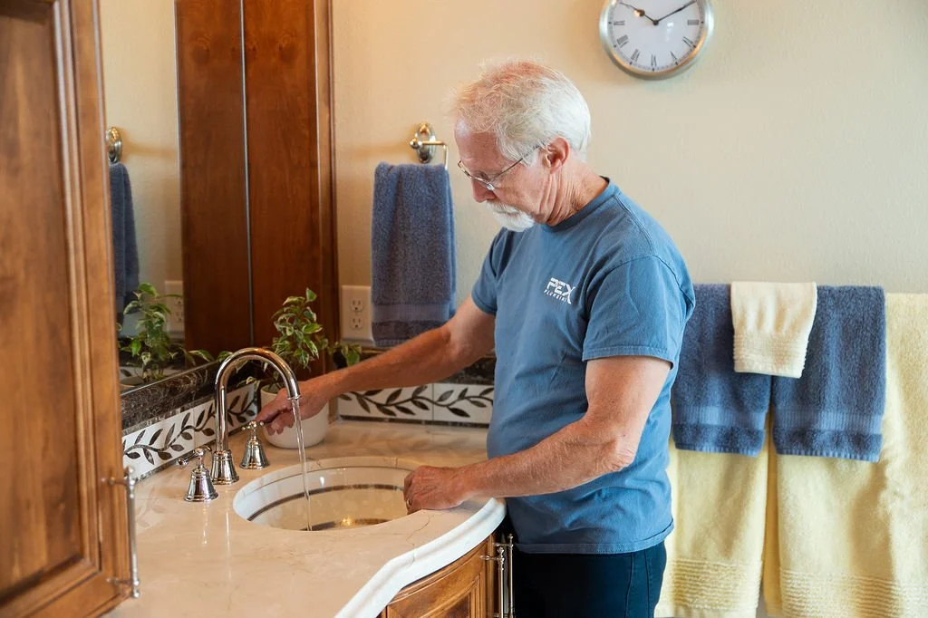 An elderly man with white hair and glasses washing his hands at a bathroom sink.