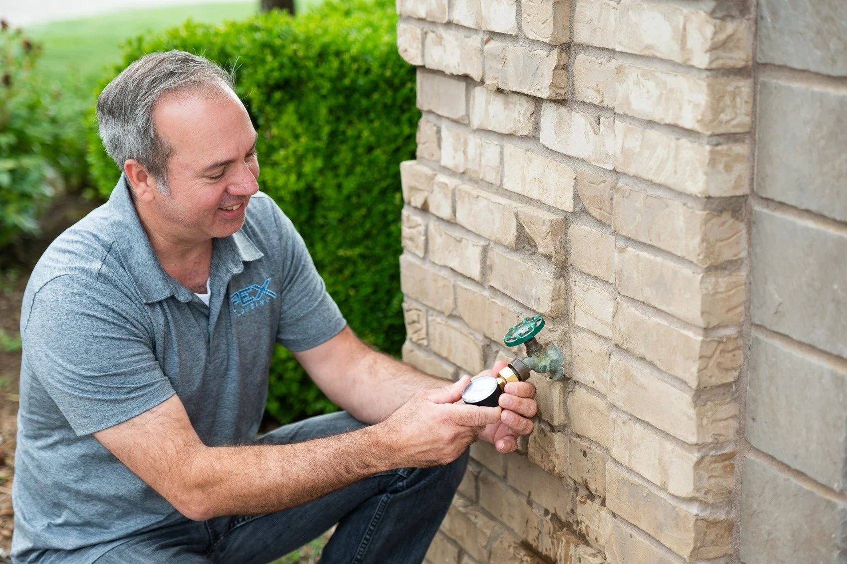 A man is adjusting a water hose faucet on a brick wall outside a house.