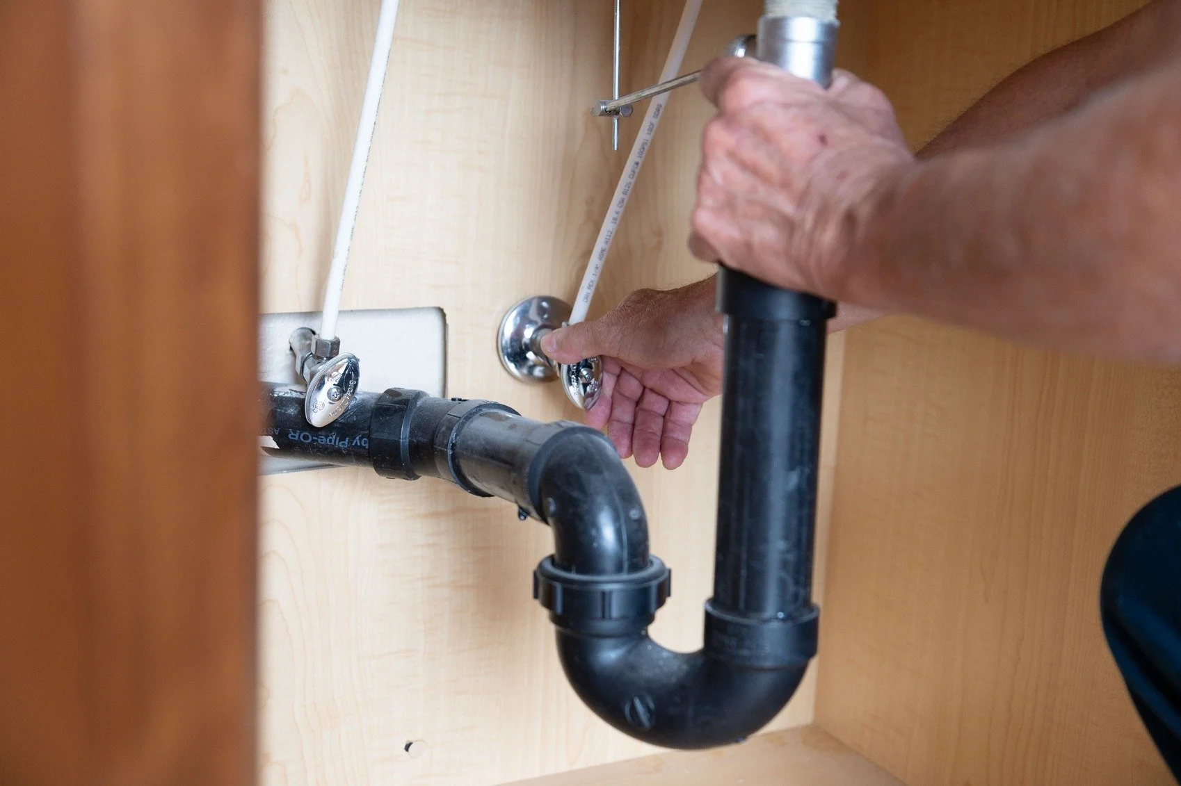 Someone using a wrench to tighten a pipe under a sink in a wooden cabinet.
