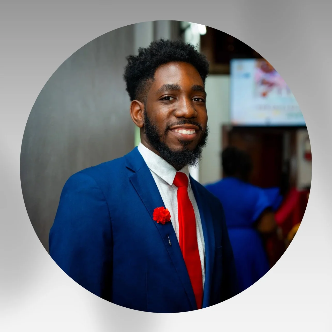 A smiling young man with dark skin, curly hair, and a beard, wearing a blue suit, white shirt, and red tie, standing indoors at a formal event.