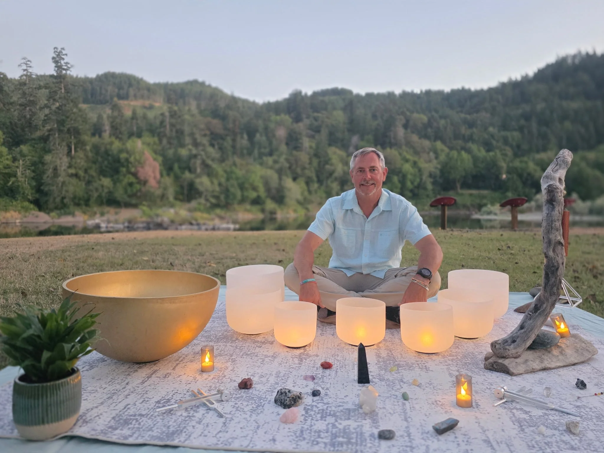 A man sits cross-legged on a white blanket outdoors at sunset, surrounded by glowing candles and crystals, with a scenic forested hillside in the background.