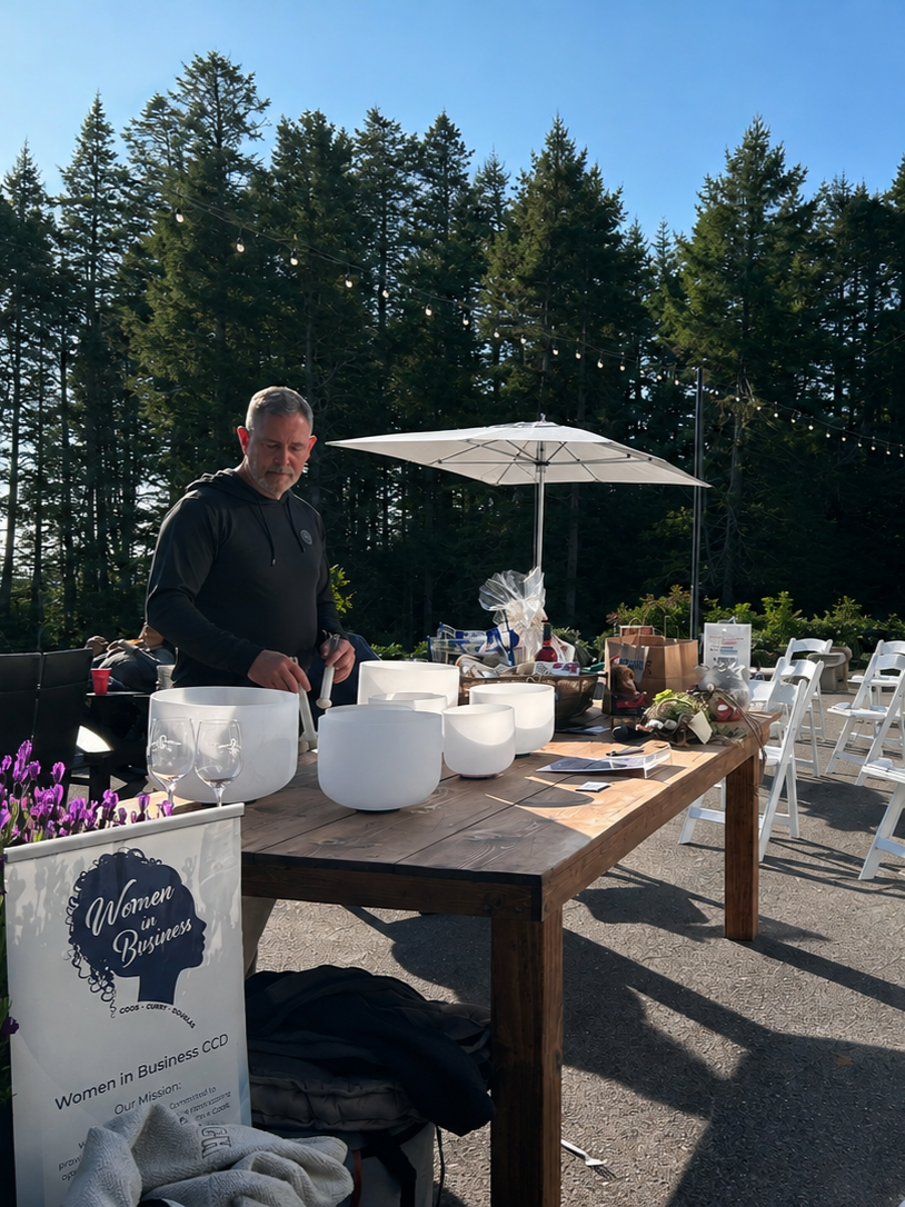 A man standing at a wooden table outdoors with crystal singing bowls, surrounded by chairs, umbrellas, and forest trees in the background.