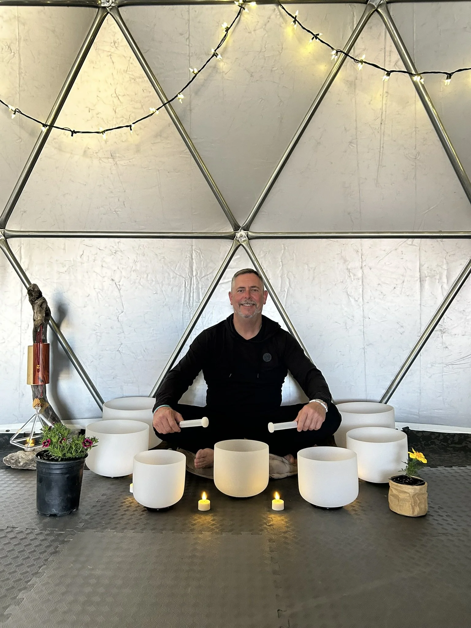 A man sitting in a seated meditation pose with a smile, surrounded by white singing bowls, candles, and plants, inside a geometric metal structure with string lights overhead.