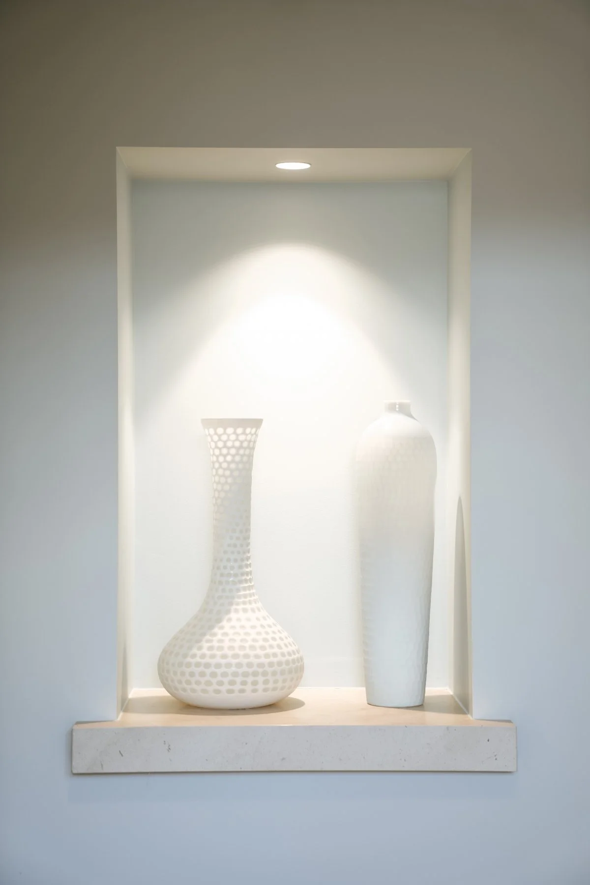 Two white vases, one with a perforated pattern and a tall, slender shape, and the other smooth and taller, displayed in a wall niche with a light above.