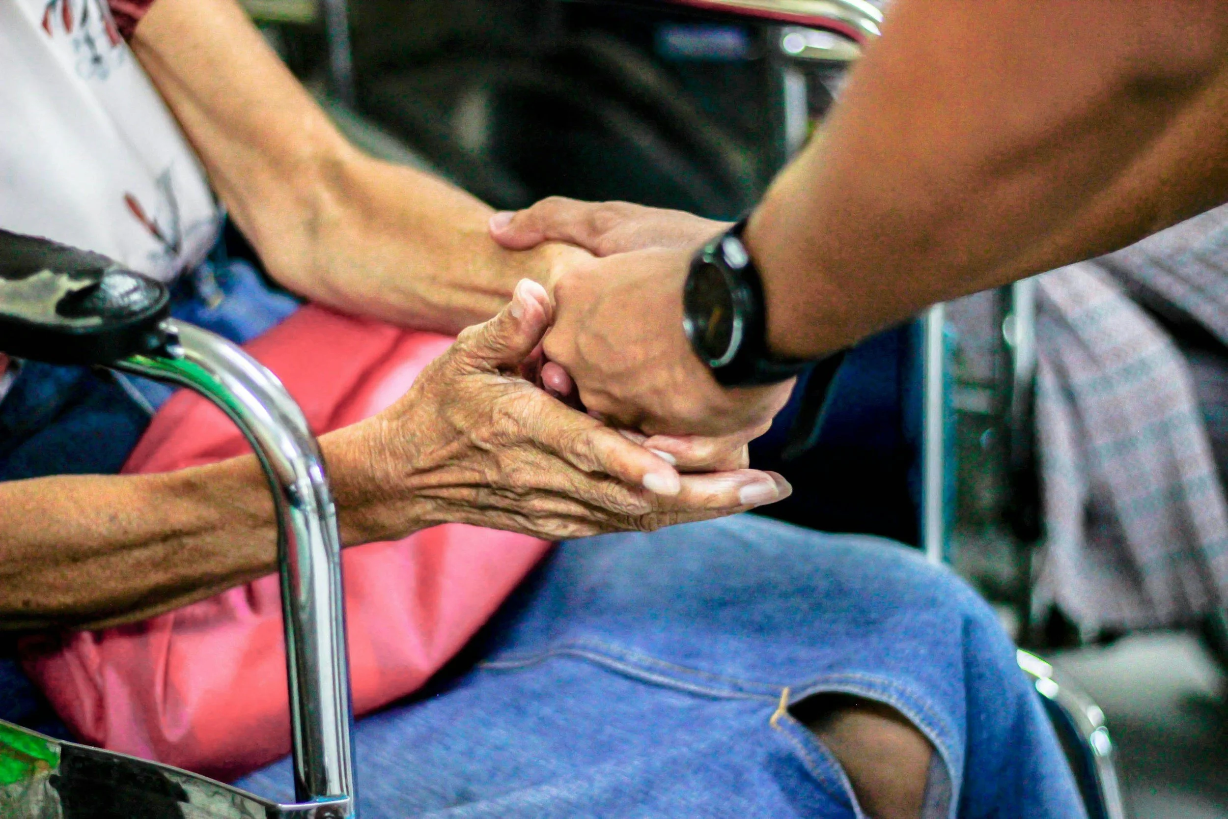 A person in a wheelchair is holding an elderly person's hand, and a younger individual is gently holding and comforting the elderly person's hand.