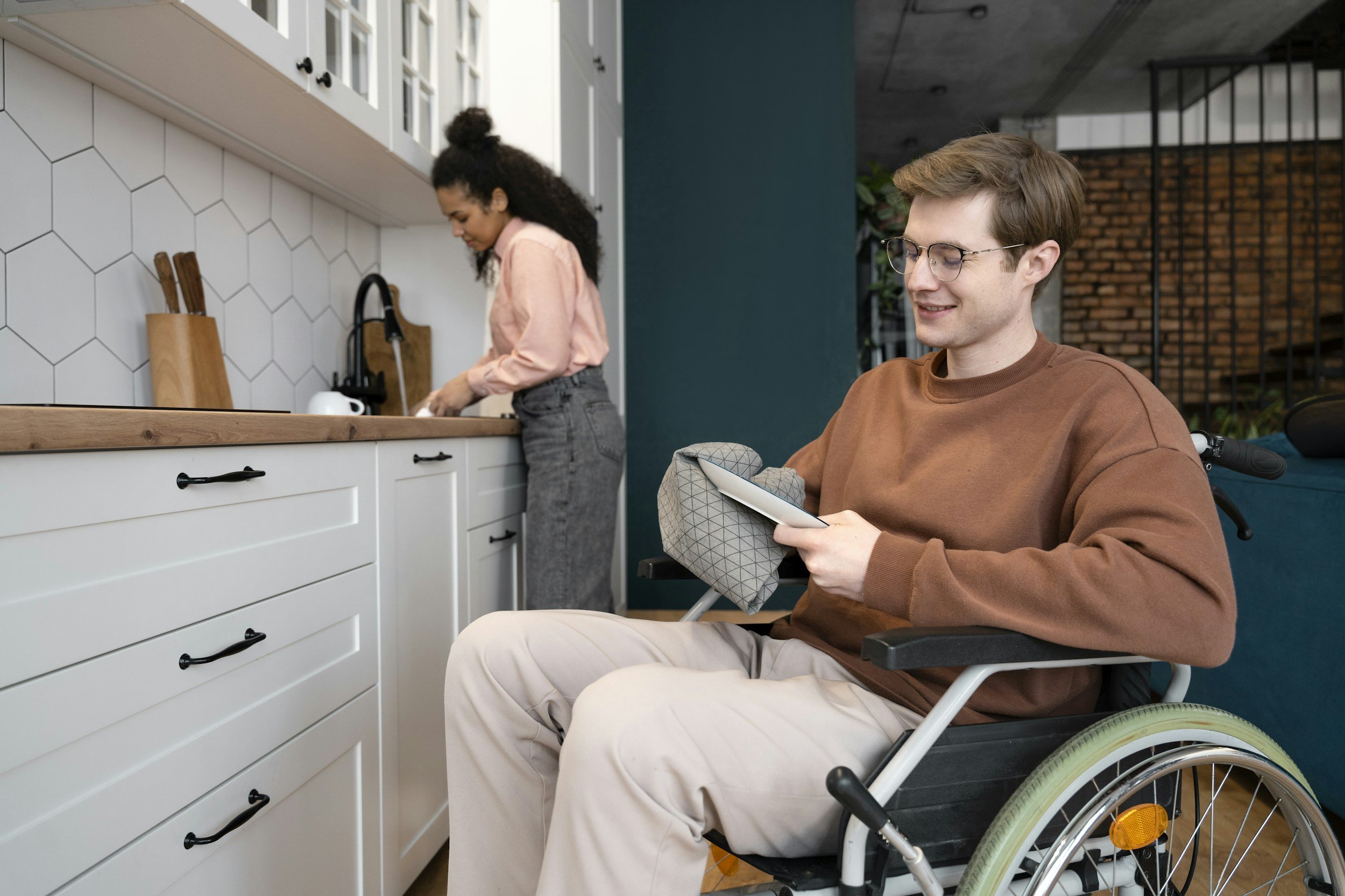 A man in a wheelchair reading a book in a modern kitchen while a woman prepares food at the counter.