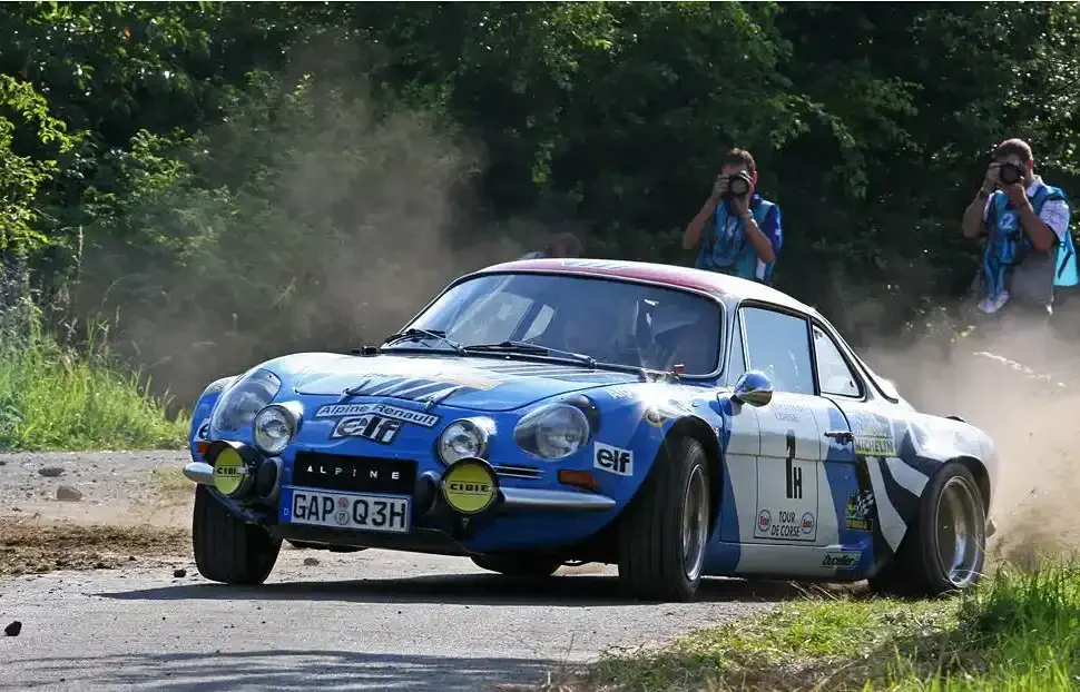 A vintage blue Alpine Renault rally car racing on a dirt road surrounded by trees, with dust and gravel kicked up, two photographers taking pictures in the background.