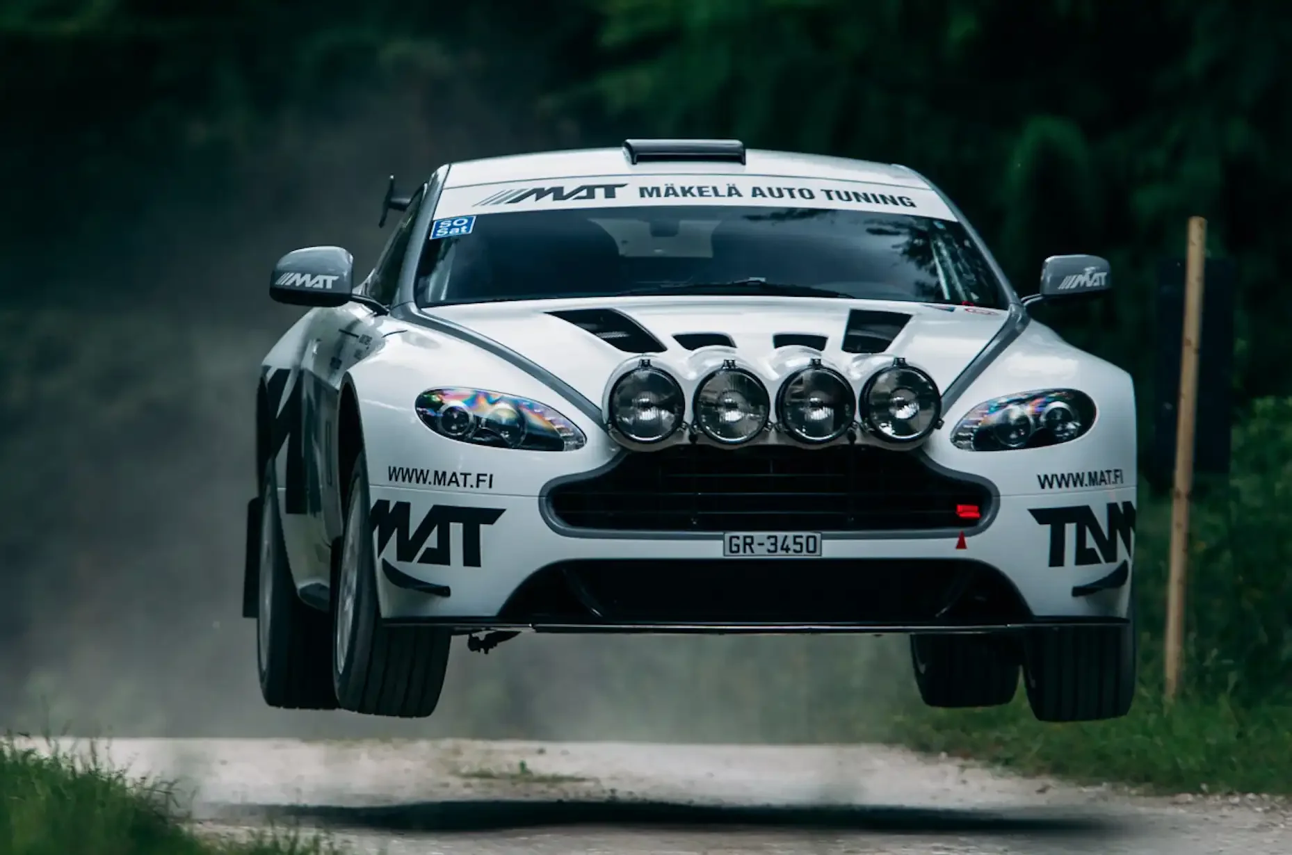 A white rally car with sponsor decals, airborne on gravel road, with four front-mounted spotlights.