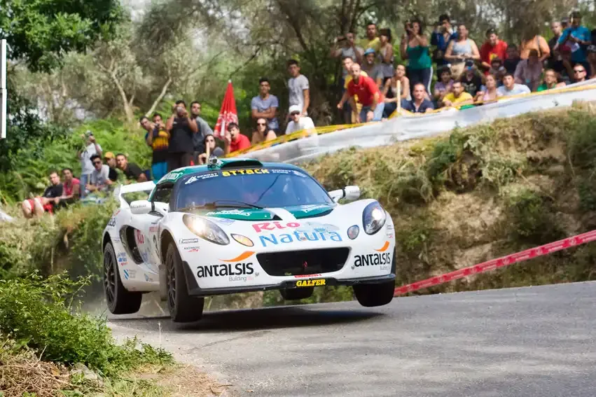 A Rally cartoon illustration, a white rally car with sponsorship decals, including 'RCT Natura' and 'ANTALISIS,' airborne during a race on a curving mountain road with a crowd of spectators watching from the roadside.