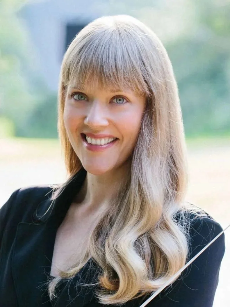 A woman with long blonde hair and blue eyes smiling outdoors in a black top.