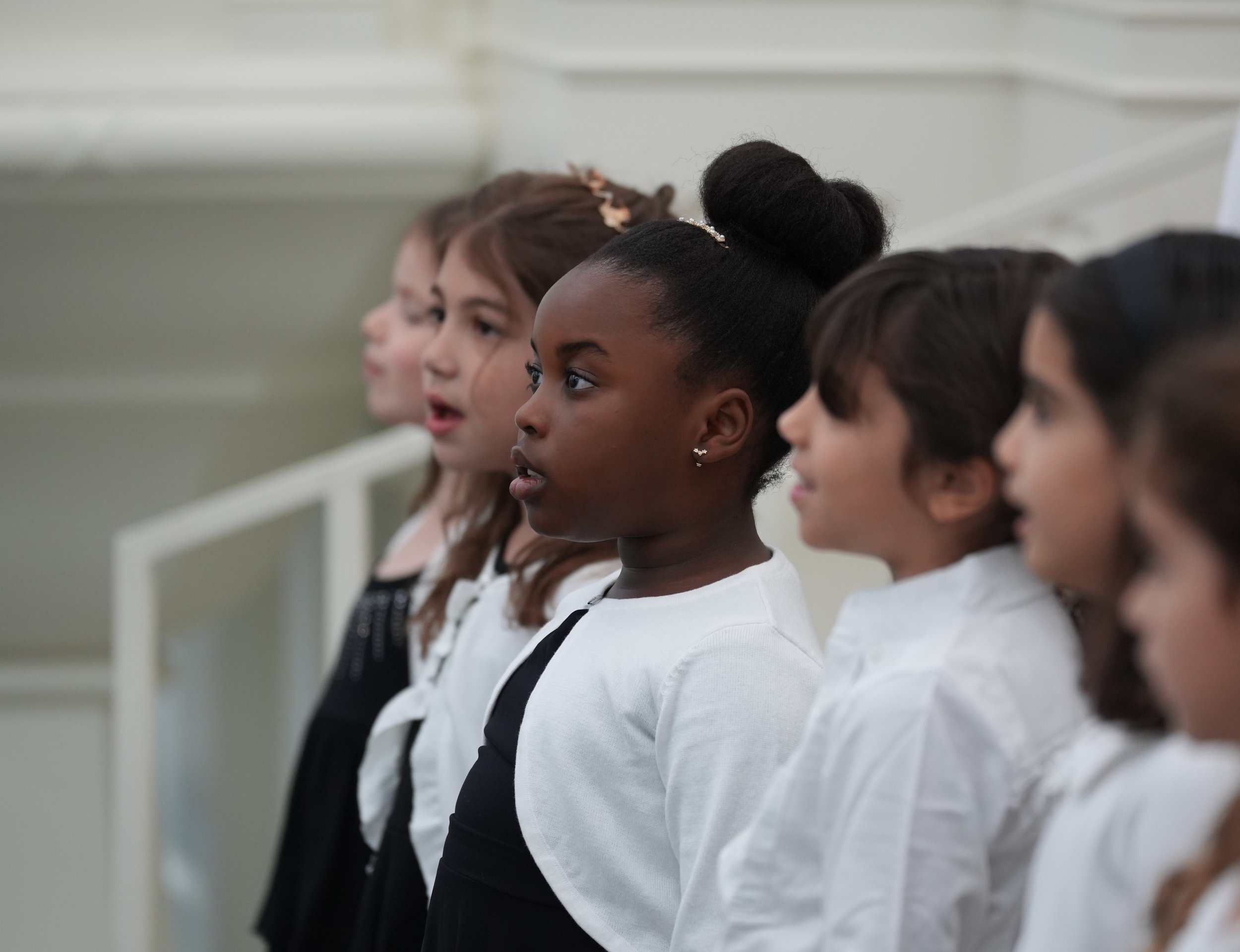 Group of young girls singing or performing in a choir, dressed in black and white outfits, standing in a row indoors.