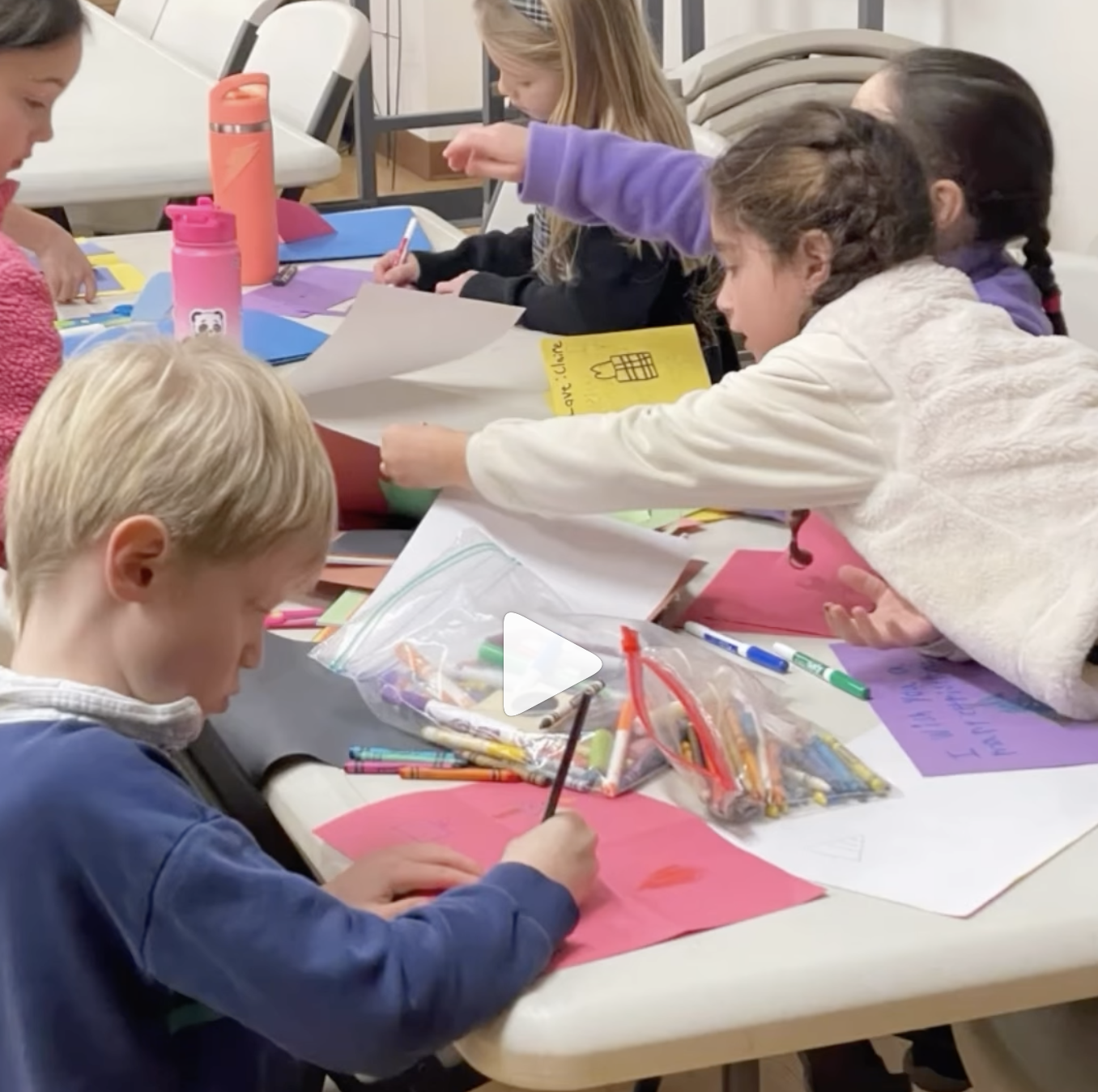 Children working on arts and crafts projects at a table with paper, markers, and supplies.