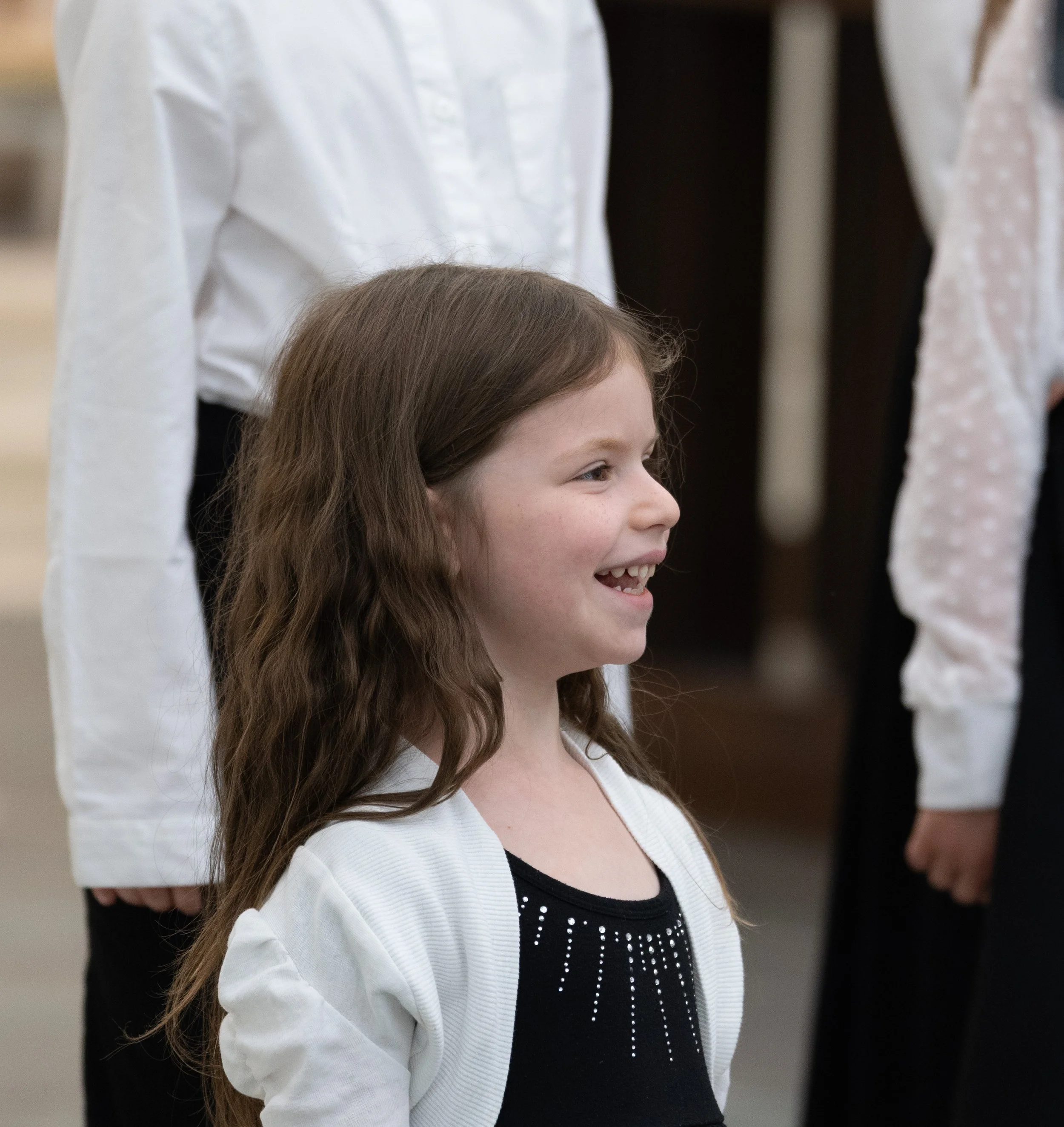 A young girl with long brown hair smiling, standing indoors with other people around her.