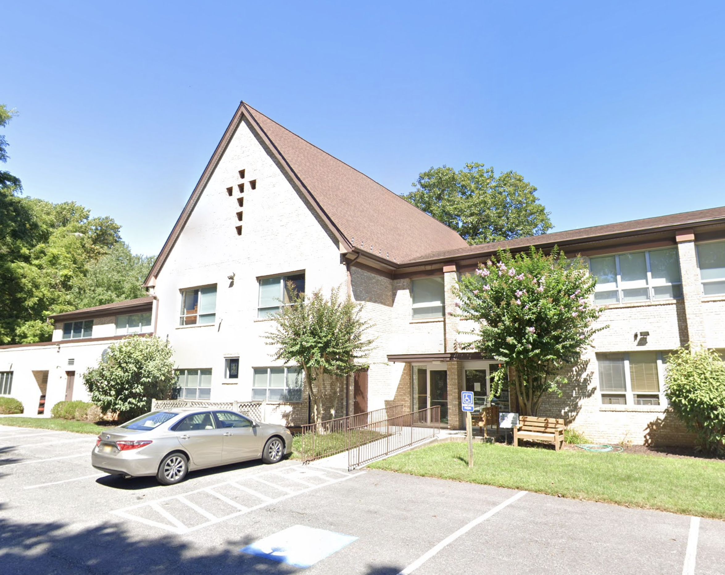 A beige brick building with a steep triangular roof and multiple windows, surrounded by trees and a parking lot with a beige sedan car.