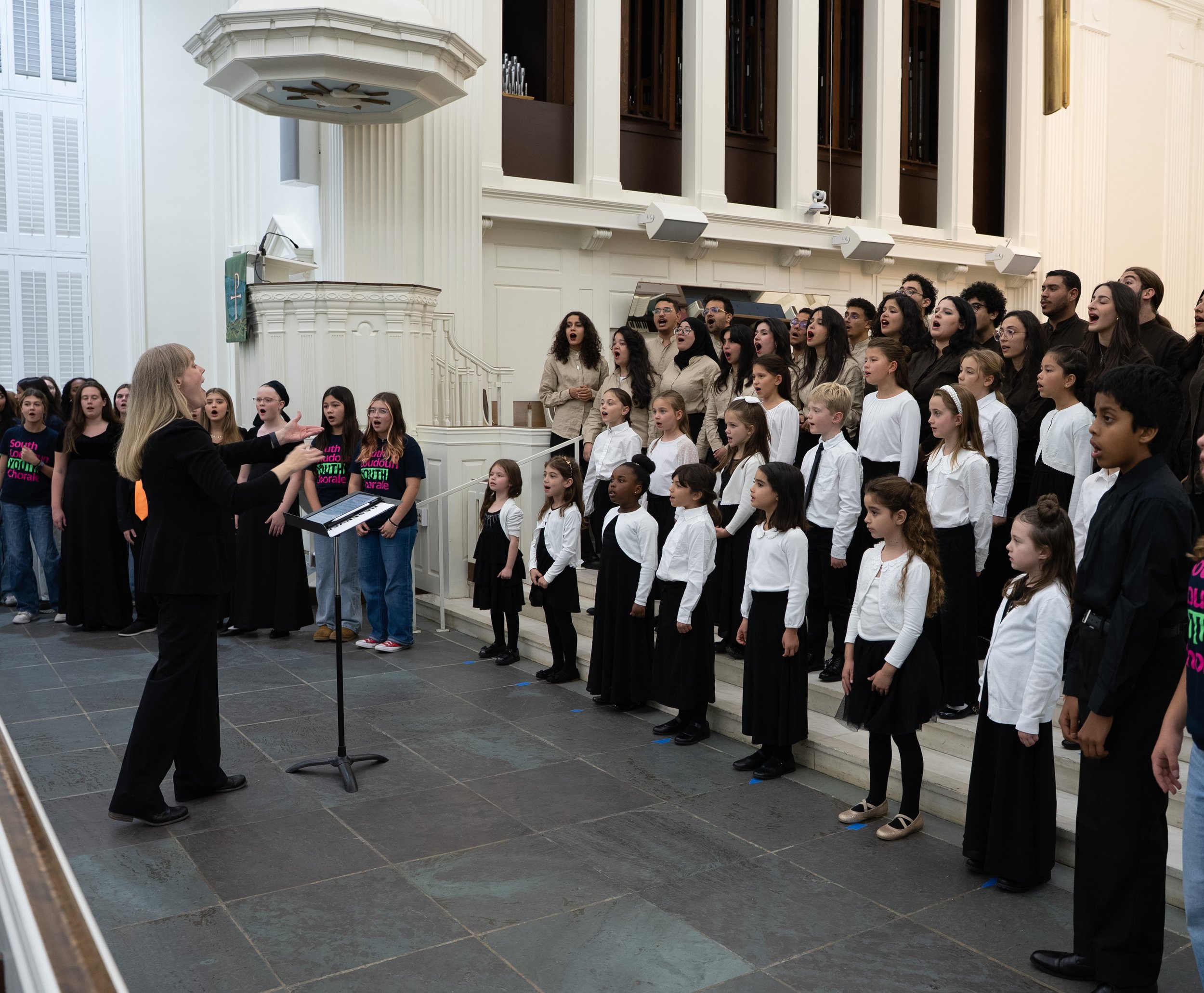A choir with children and adults singing during a performance in a church, directed by a female conductor.