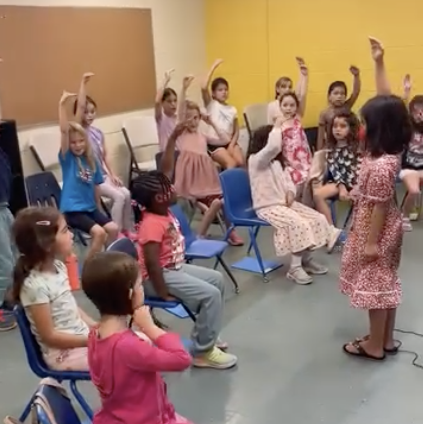 A group of young children, seated on chairs, some raising their hands, in a classroom or activity room, with a woman standing in front of them.