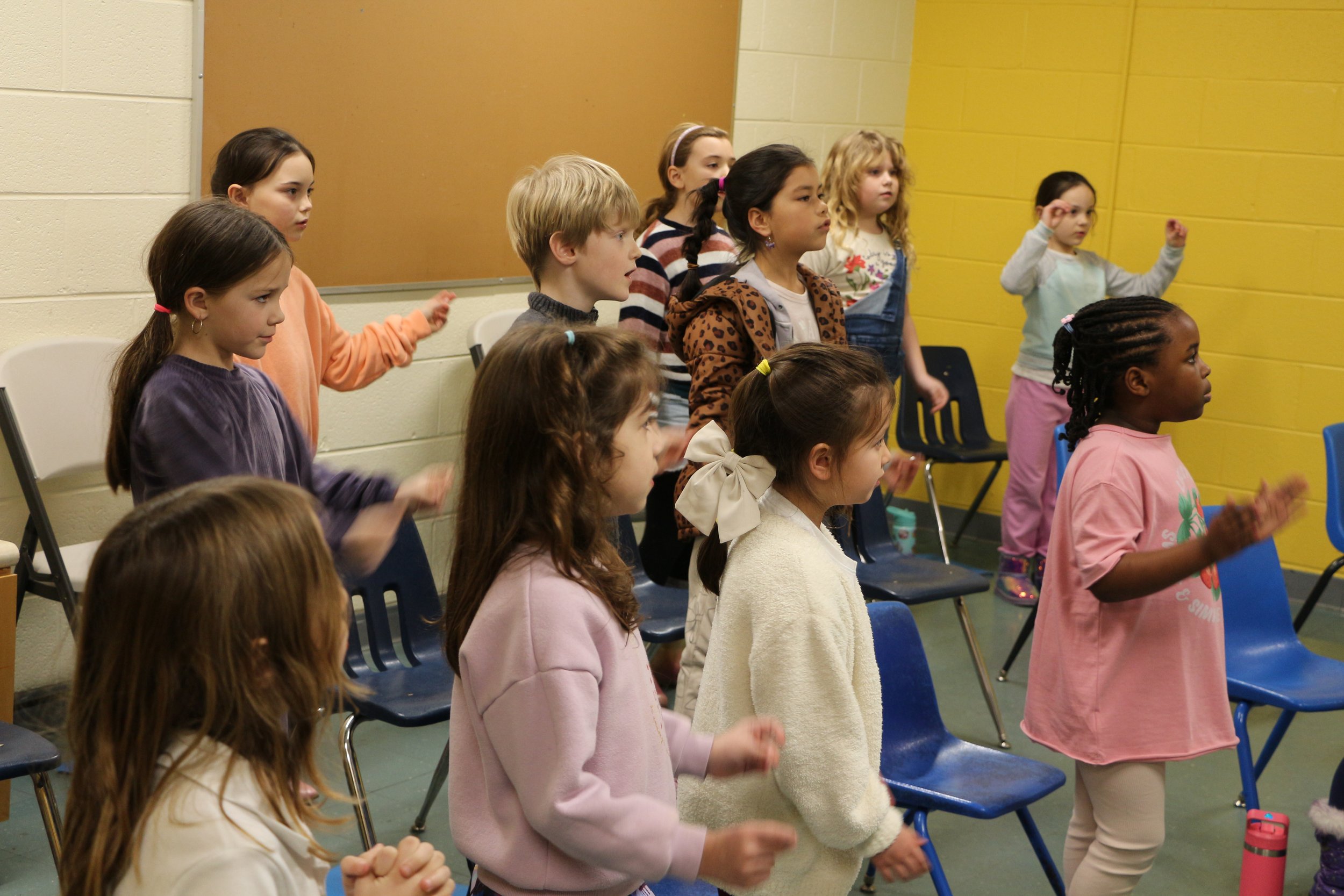 Group of children standing and participating in an activity in a classroom, some have hands raised or clapping, with chairs lined up against the wall.