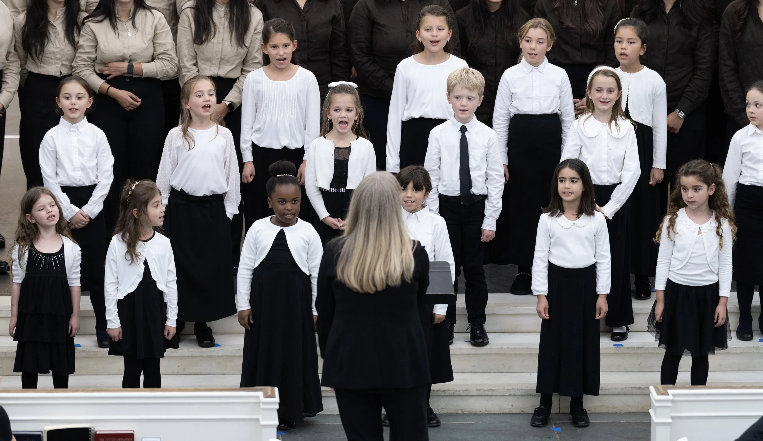Children dressed in white and black performing in a choir with a woman conducting on stage.