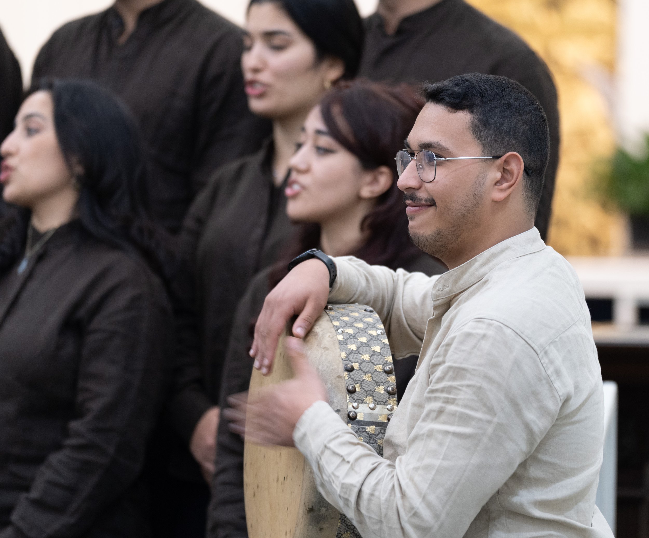 A group of five people, dressed in black, standing close together with one man in the foreground playing a hand drum and smiling.