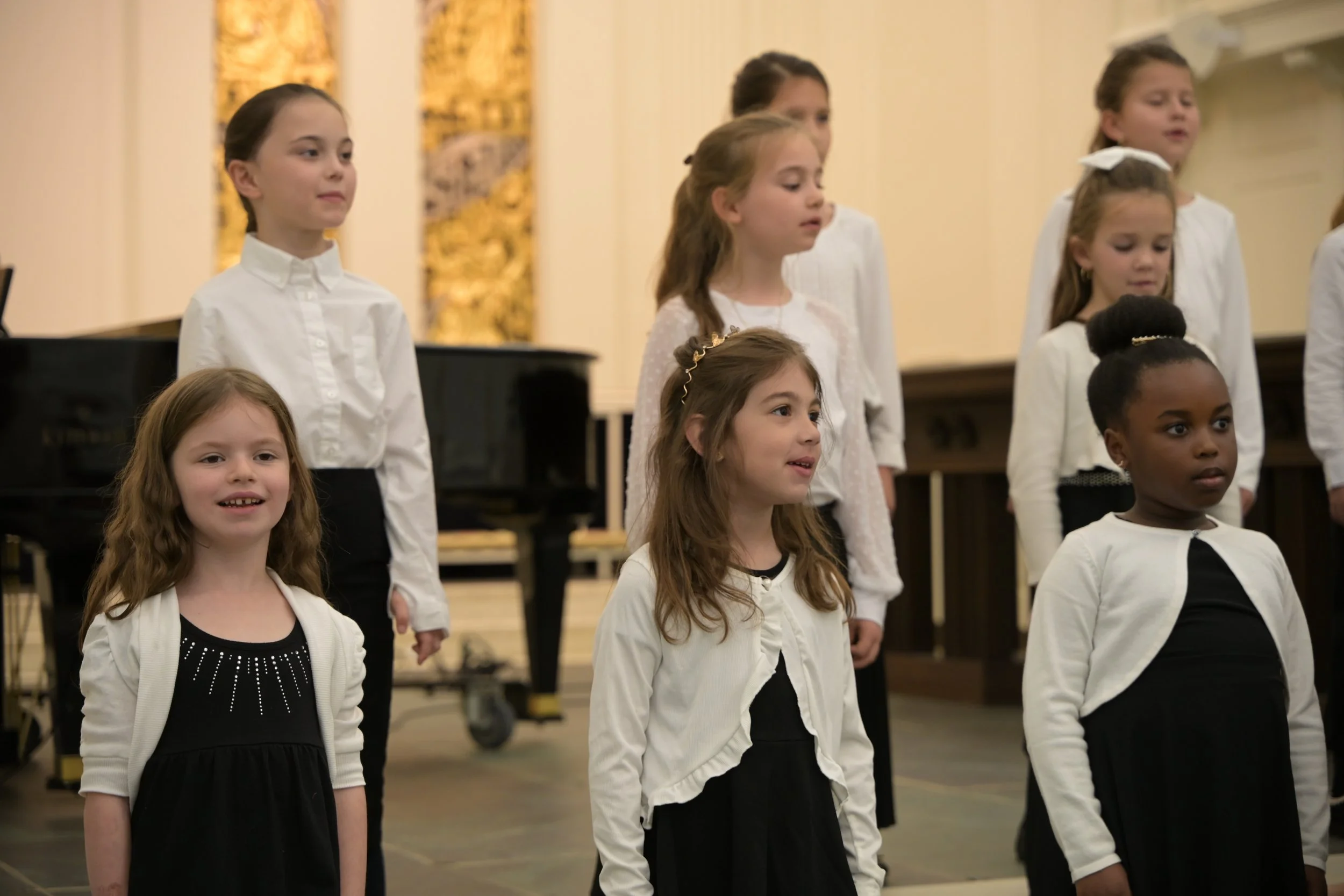 Young girls singing in a choir, dressed in black and white, standing in rows in a formal setting with a grand piano in the background.