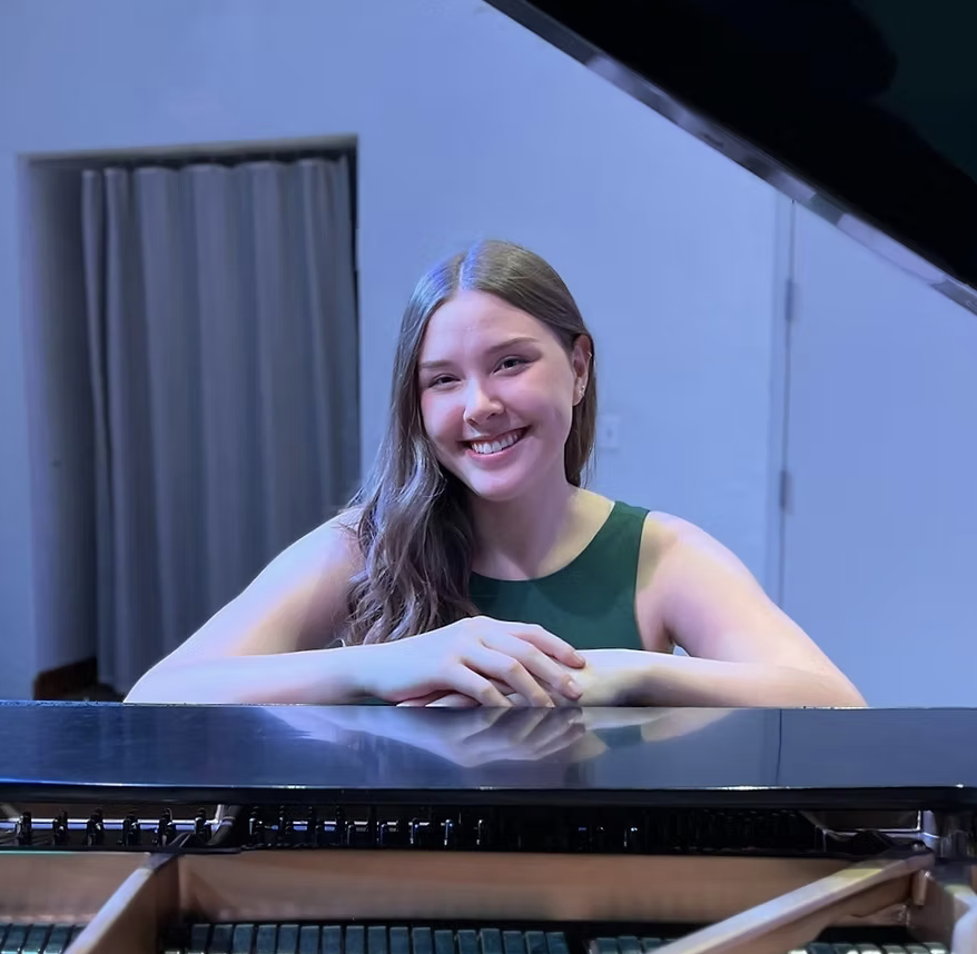 Young woman with long brown hair smiling while sitting at a piano with the lid open, in a room with gray walls and a window with curtains.