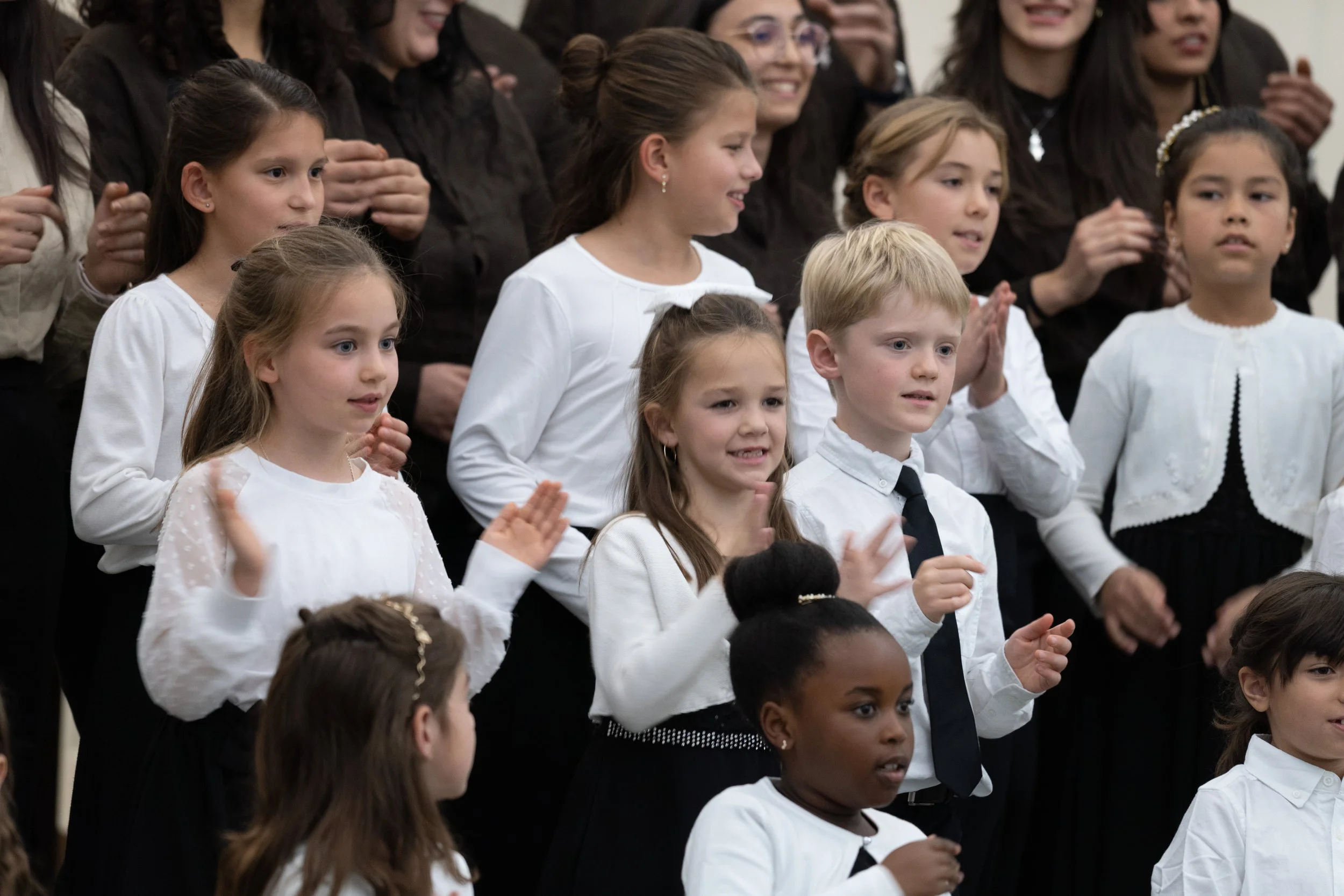 Children dressed in white and black choir robes singing or performing together, some with hands clapping, in a group setting.
