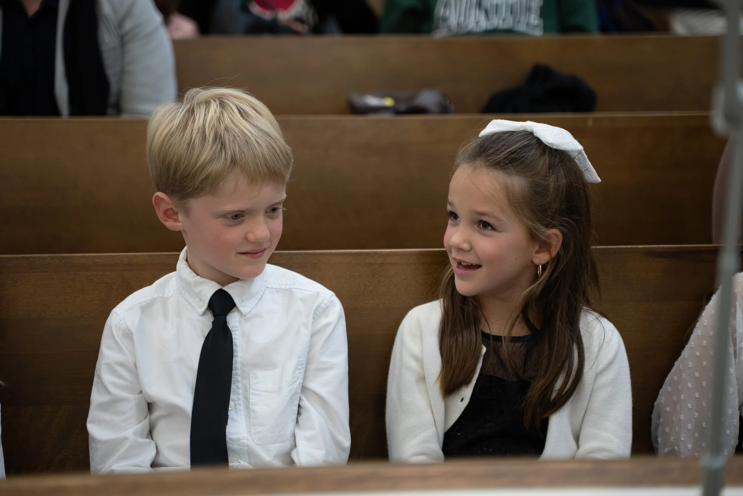 A young boy with blonde hair wearing a white shirt and black tie, sitting next to a young girl with brown hair, white bow, and earrings, both smiling and talking in a church pew.