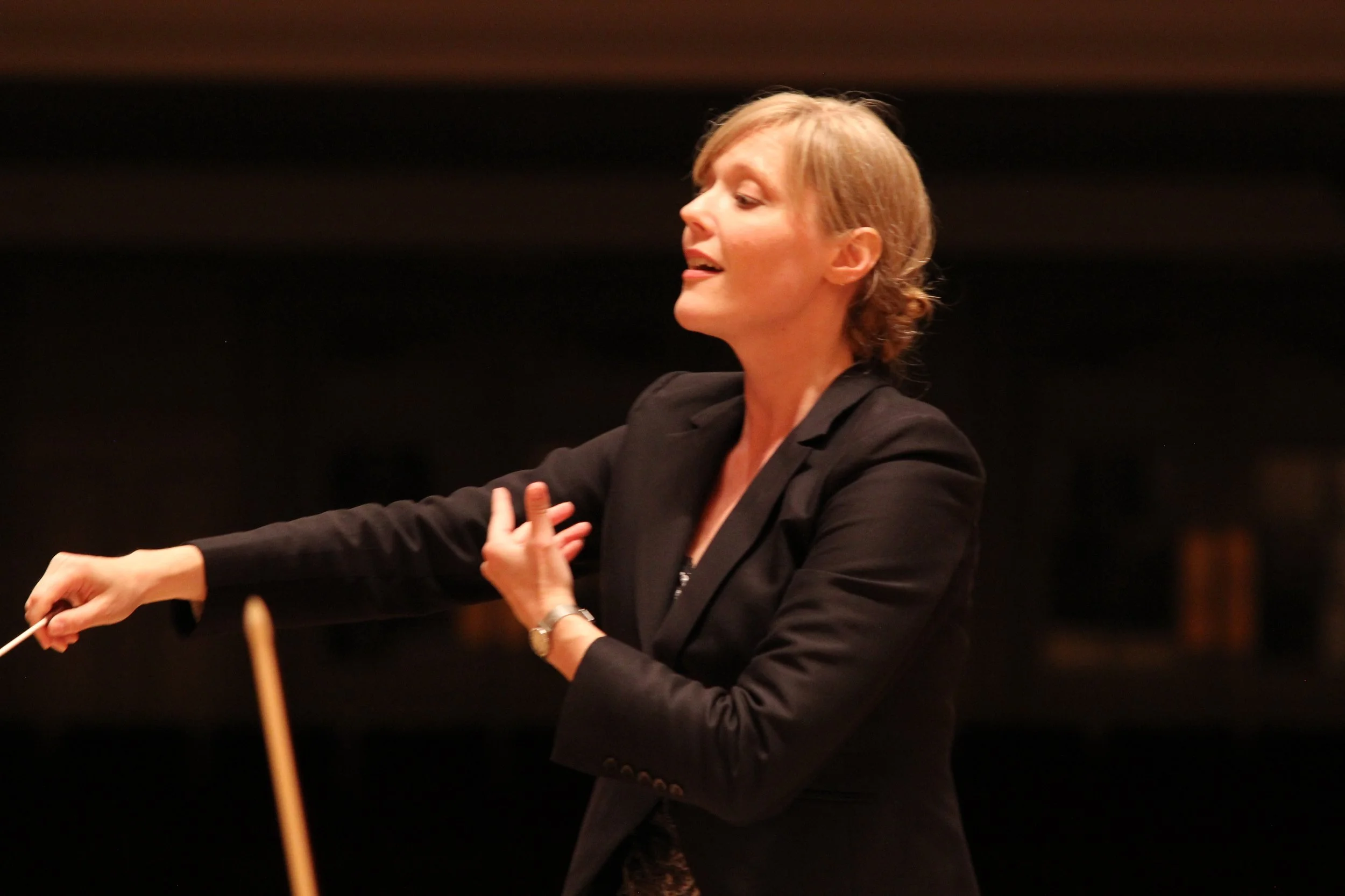 A woman in a black blazer conducting an orchestra with a baton in her right hand.