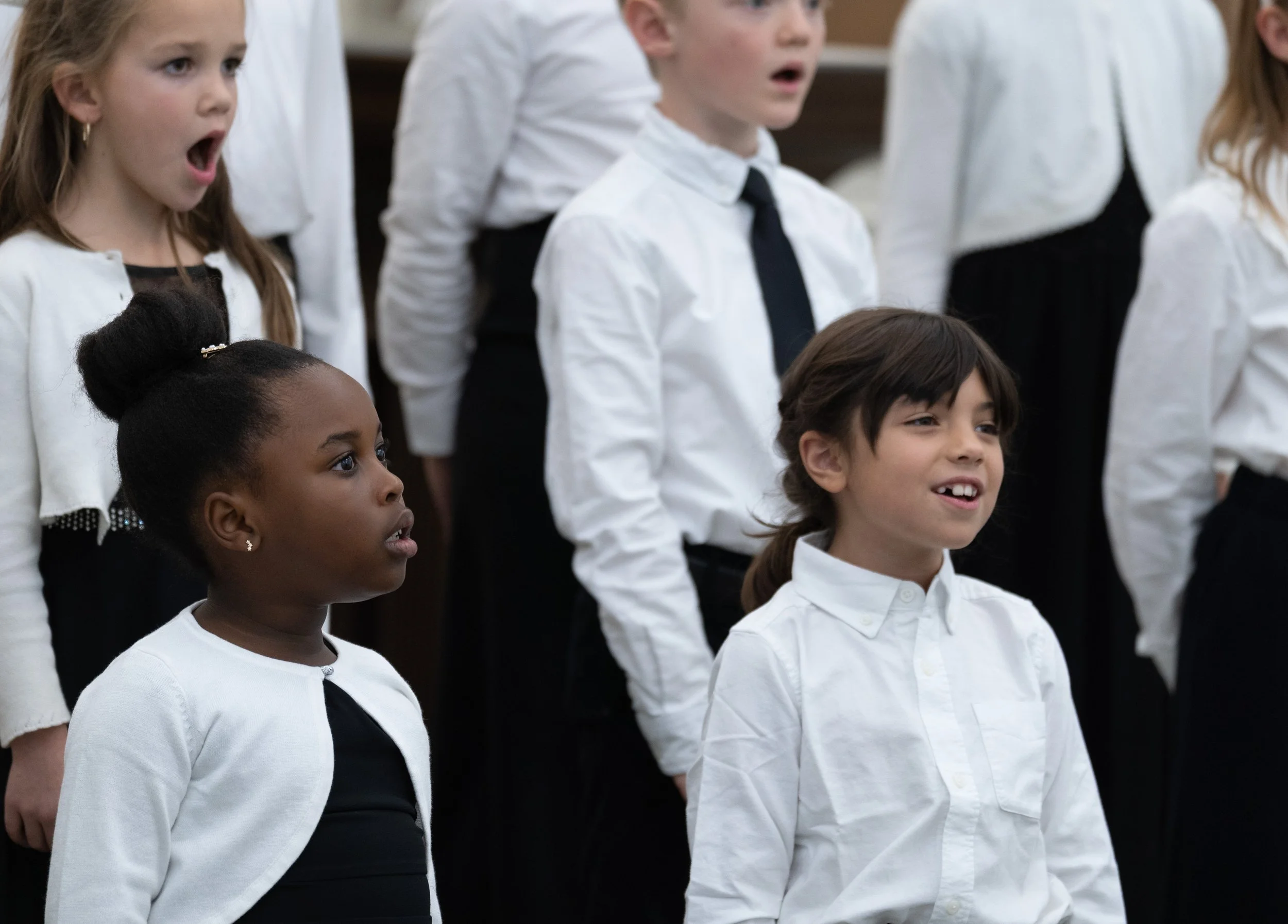 Children in a choir singing, dressed in white shirts and black bottoms, with expressions of concentration and emotion.