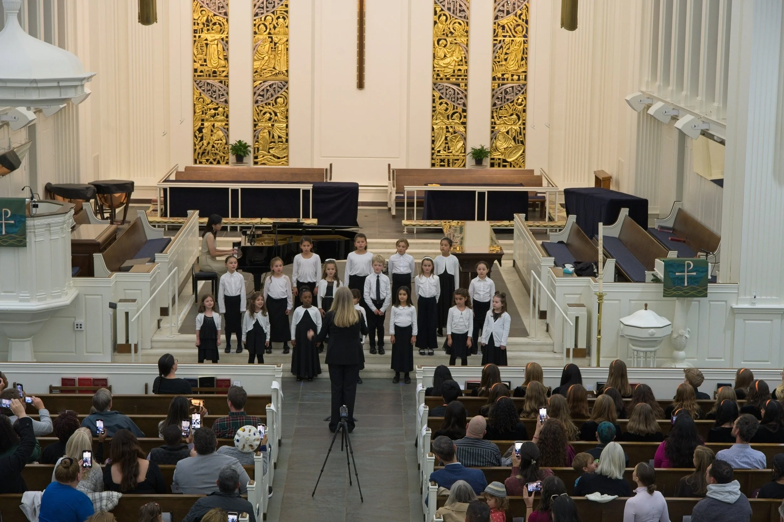 Children standing on stage in a church, singing with a conductor, with an audience seated and taking photos.
