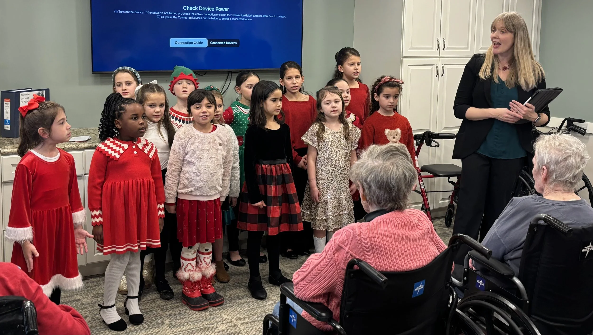 A group of young girls dressed in holiday-themed clothing, some in red dresses and festive accessories, are singing or performing for an audience of elderly people in wheelchairs. A woman stands to the right, possibly the conductor or teacher, holding a clipboard and speaking to the performers. The room has a large TV screen displaying instructions for checking device power and connected devices.