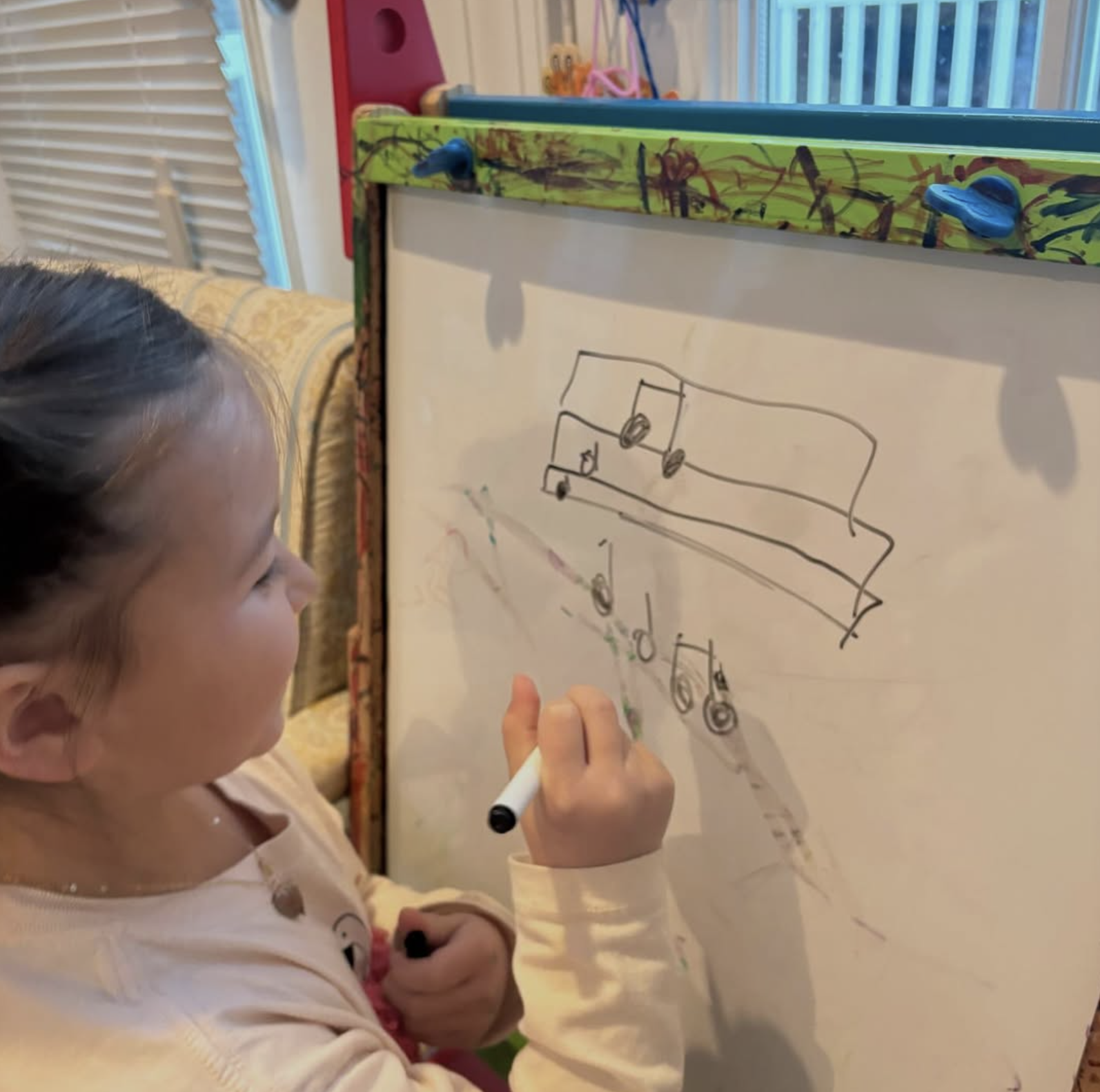 A young girl drawing on a whiteboard with a black marker. Her cowlick hairstyle is visible. The whiteboard has colorful markers and a wooden frame with paint splatters.