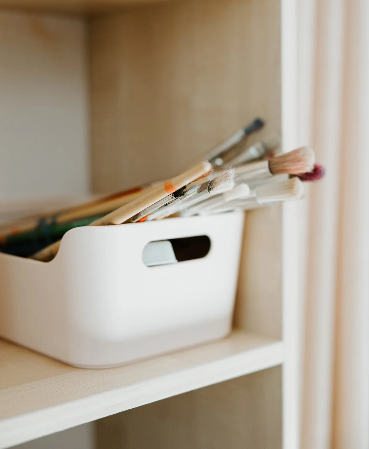 Close-up of a white storage box containing various paintbrushes, placed on a wooden shelf near a window.