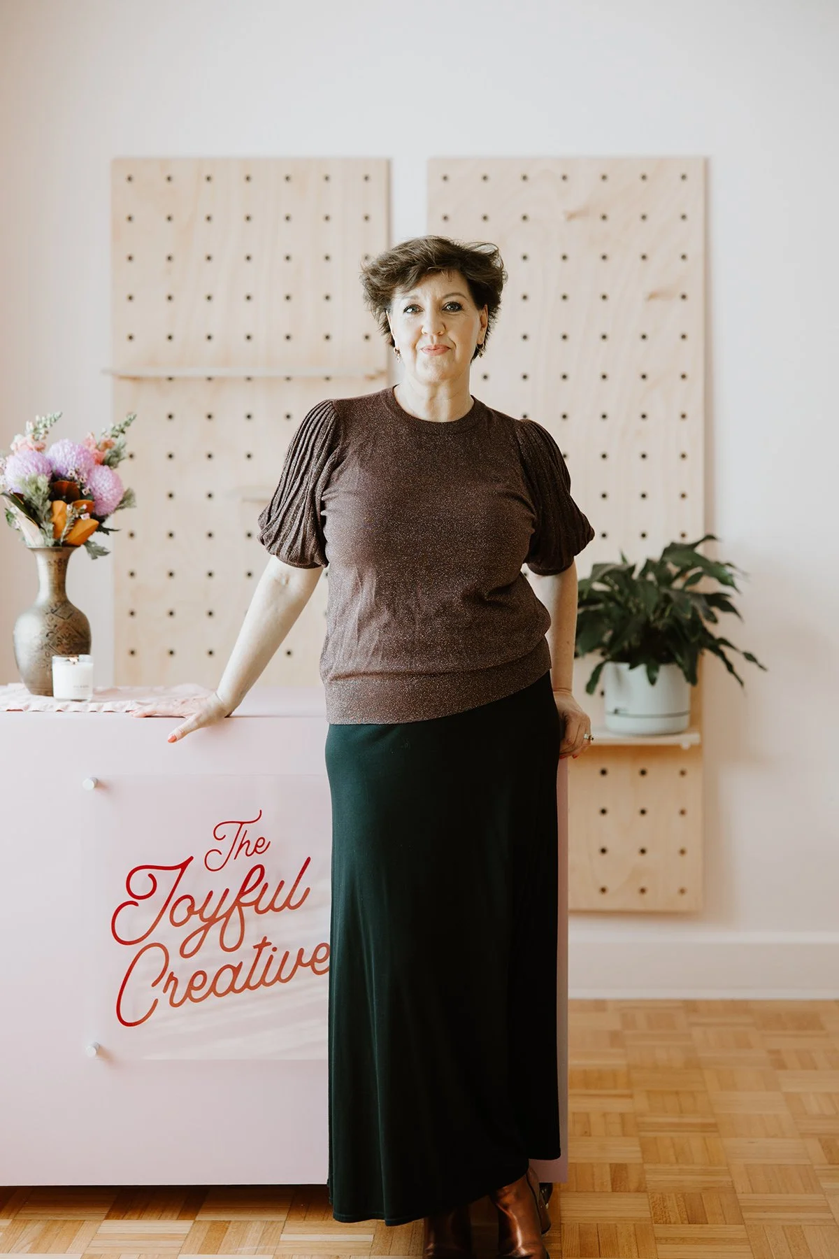 A woman standing indoors next to a pink table with a floral arrangement and a candle, with a wooden pegboard wall and potted plant in the background.