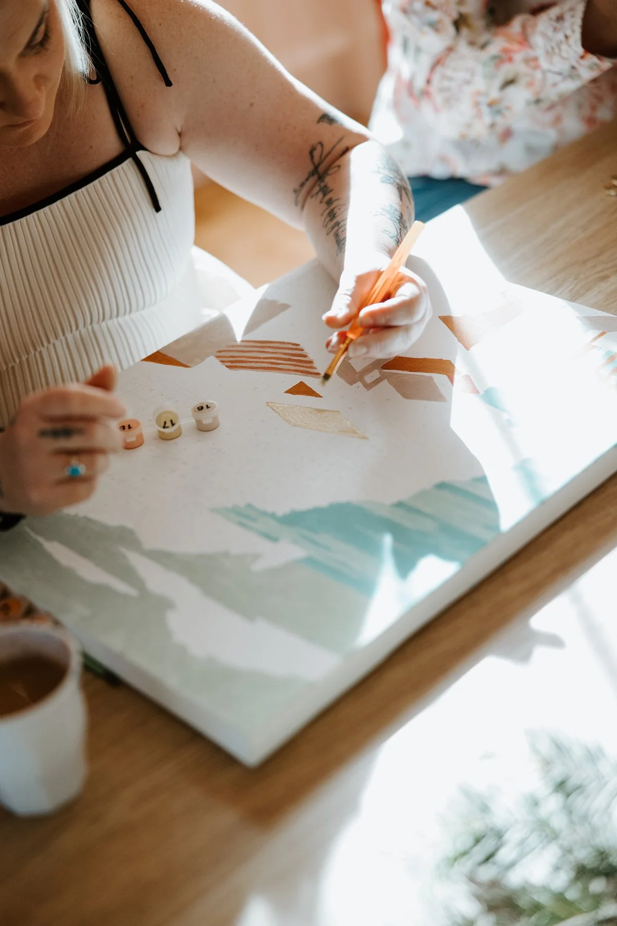 Person working on an abstract geometric painting or collage at a table, with small, numbered paint or color tokens nearby, in a well-lit room.