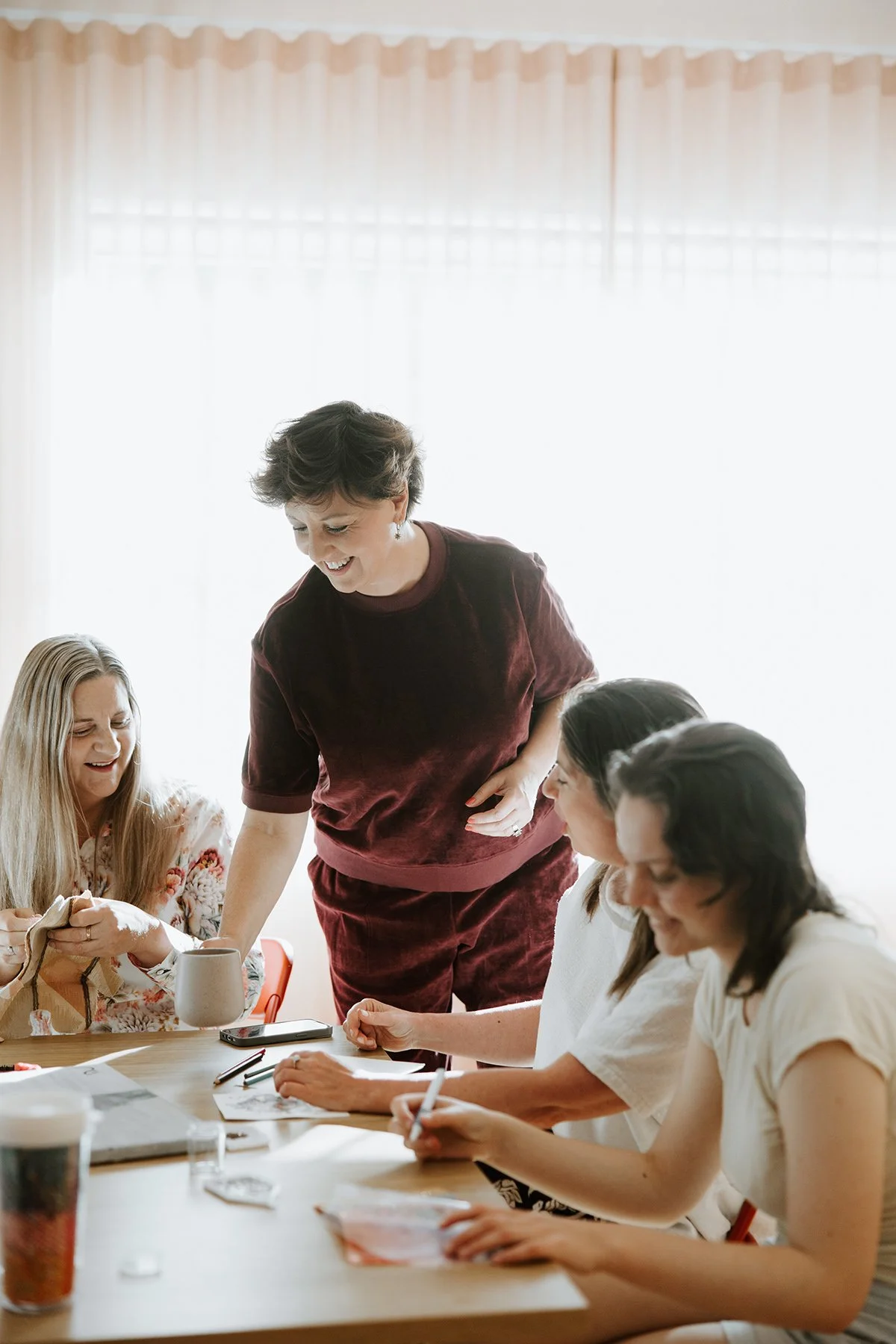A woman in a maroon outfit smiling and engaging with three women seated at a table, during a casual meeting or discussion in a bright room.