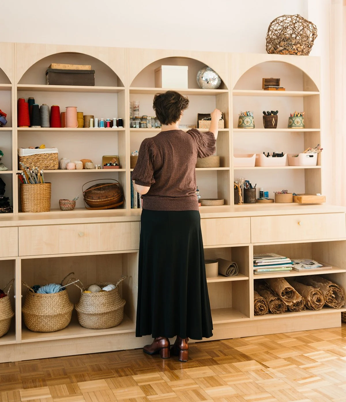 A woman standing in front of a wooden shelf filled with yarn, books, baskets, and craft supplies in a craft room.